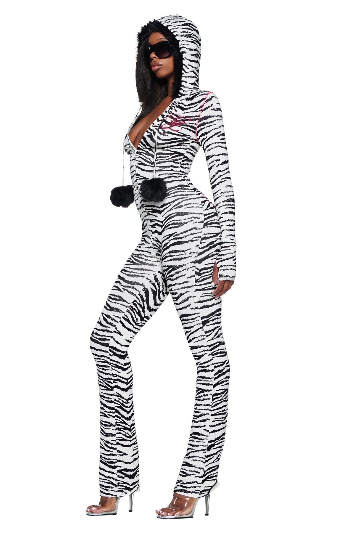 A person wears a hooded, zebra-print YVL BLARE FUR LINED JUMPSUIT - ZEBRA with a fur-trimmed hood and pom-pom drawstrings, paired with clear high-heeled sandals.