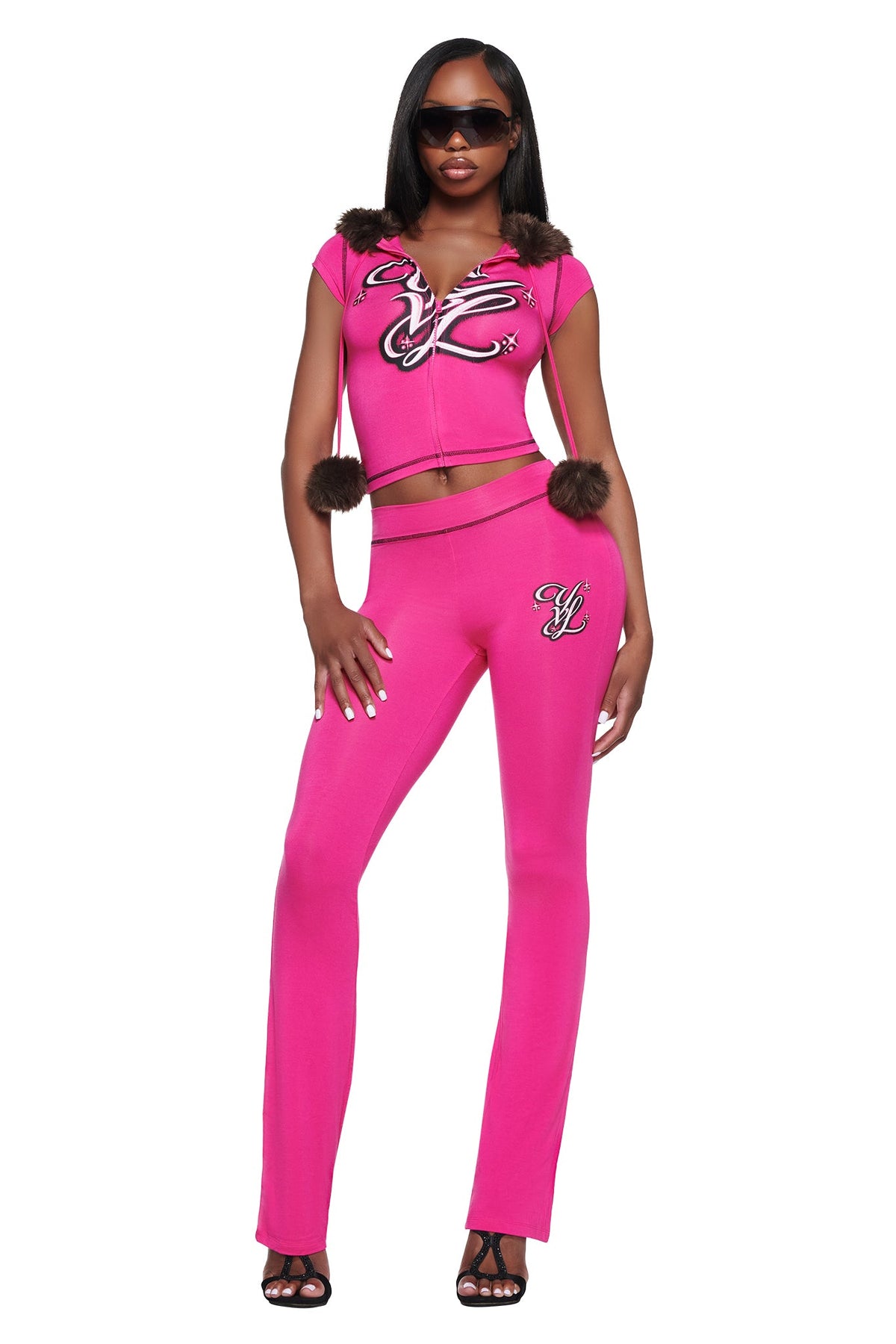 A person wearing a bright pink zip-up hoodie with brown fur trim and matching YVL BLARE TRACKPANT - PINK with a flared leg and embroidered logos.