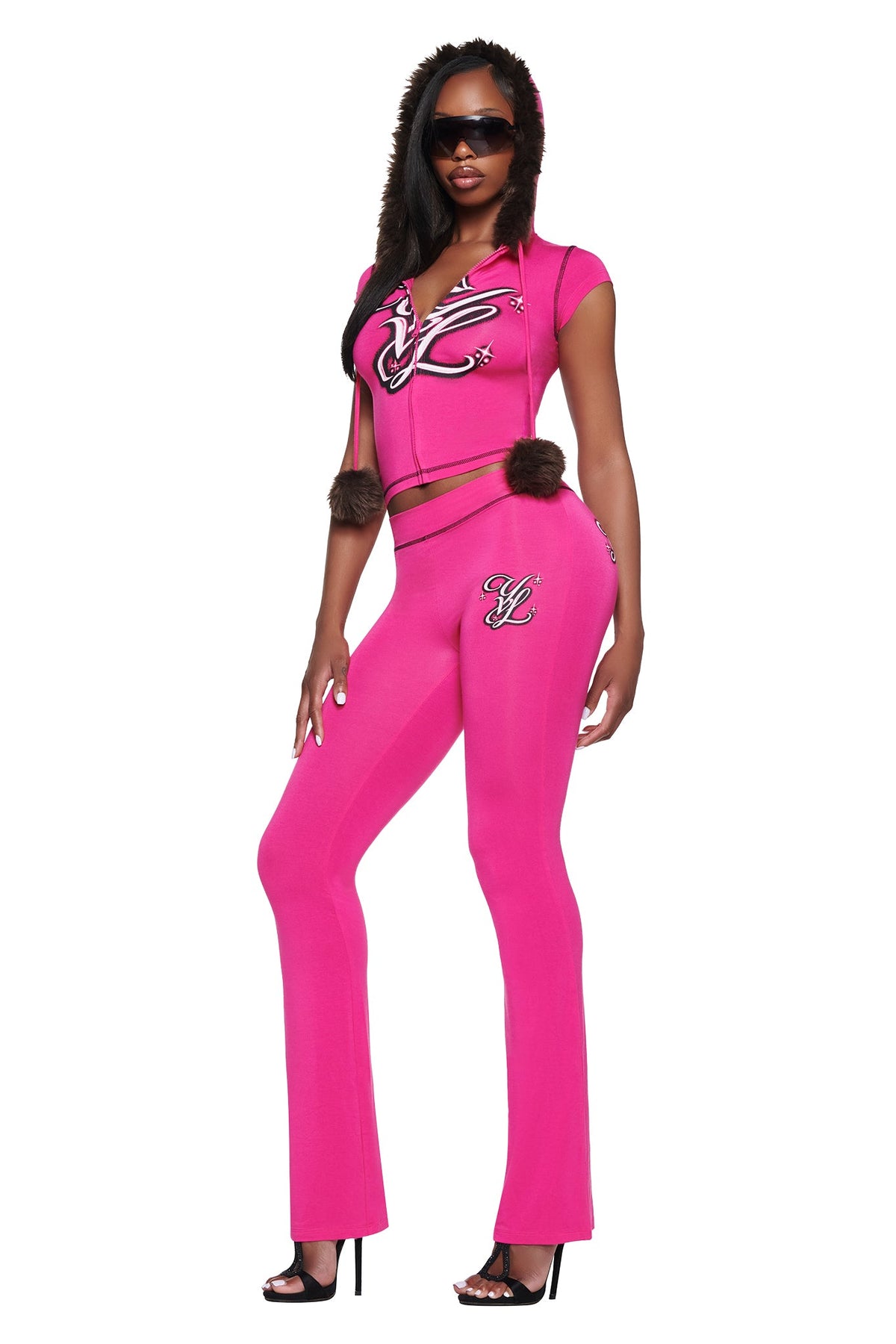 A person wears a matching hot pink hooded zip-up crop top with faux-fur trim and flared YVL BLARE TRACKPANT - PINK, both featuring embroidered logo detailing.