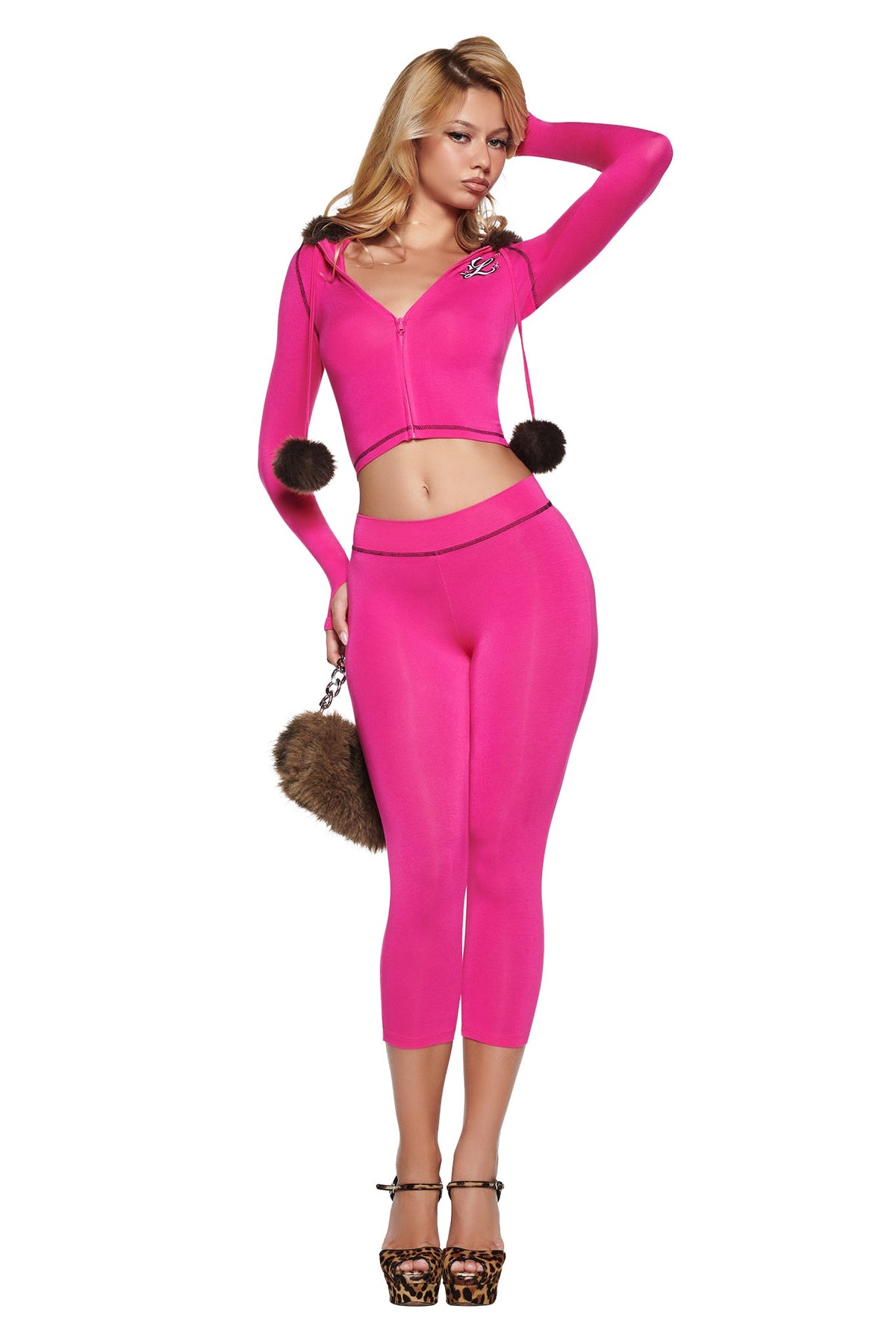 A woman wearing an I.AM.GIA YVL BLARE CAPRI - PINK outfit consisting of a bright pink zip-up cropped hoodie with a dark fur-trimmed hood and pom-poms, matching capri leggings, and leopard-print heels, holding a fuzzy brown handbag.