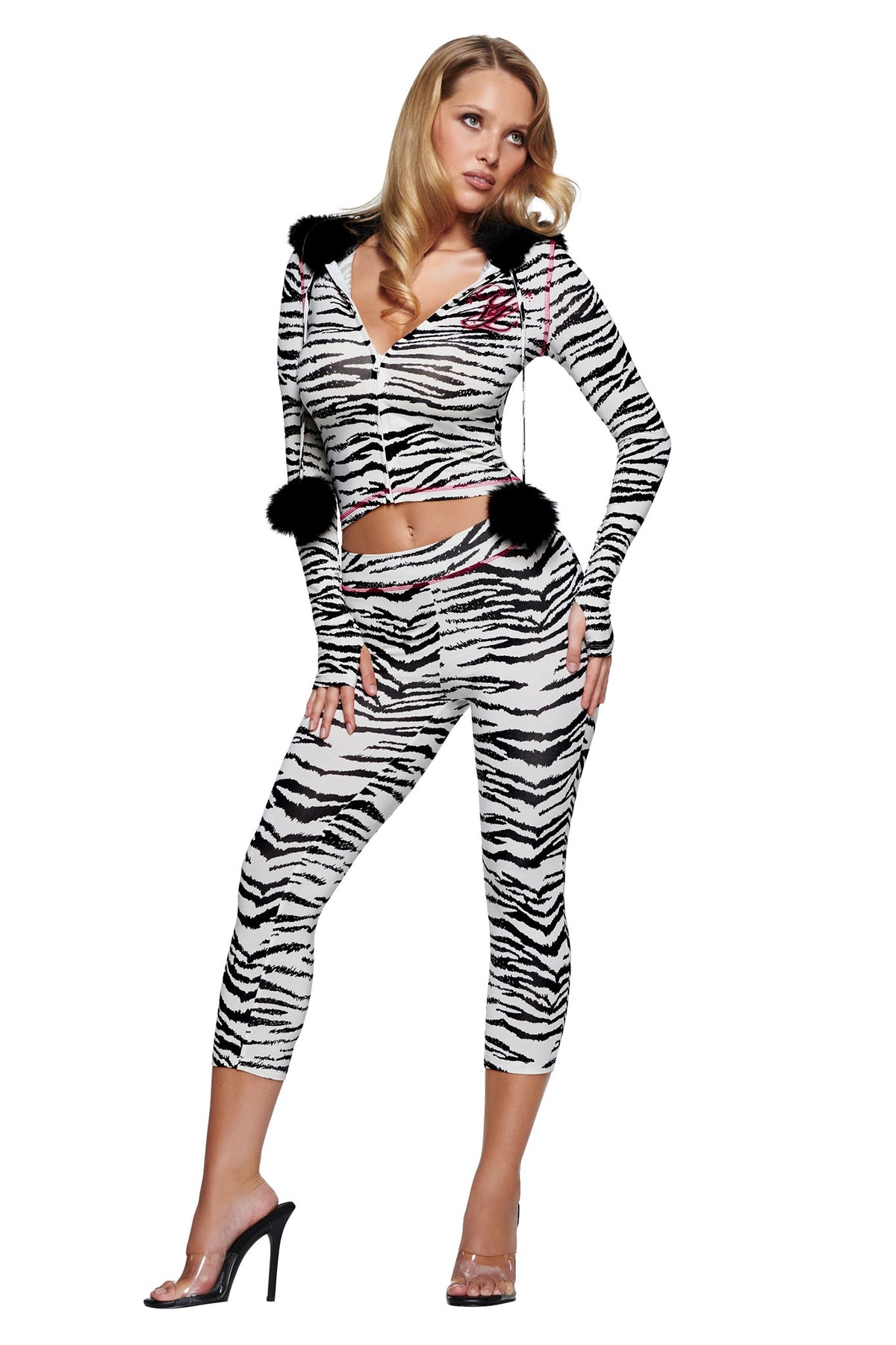 A woman wears an I.AM.GIA YVL BLARE CAPRI - ZEBRA set consisting of a zippered hoodie with black pom-poms and matching leggings, paired with clear high-heeled mules.