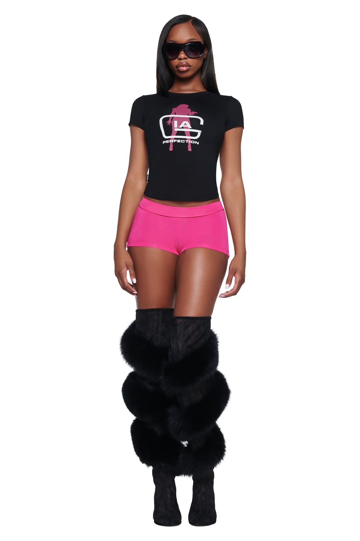 A model wears a black, short-sleeved cropped YVL T-SHIRT - BLACK GIA with a pink graphic logo, bright pink shorts, and tall black boots with faux fur trim.