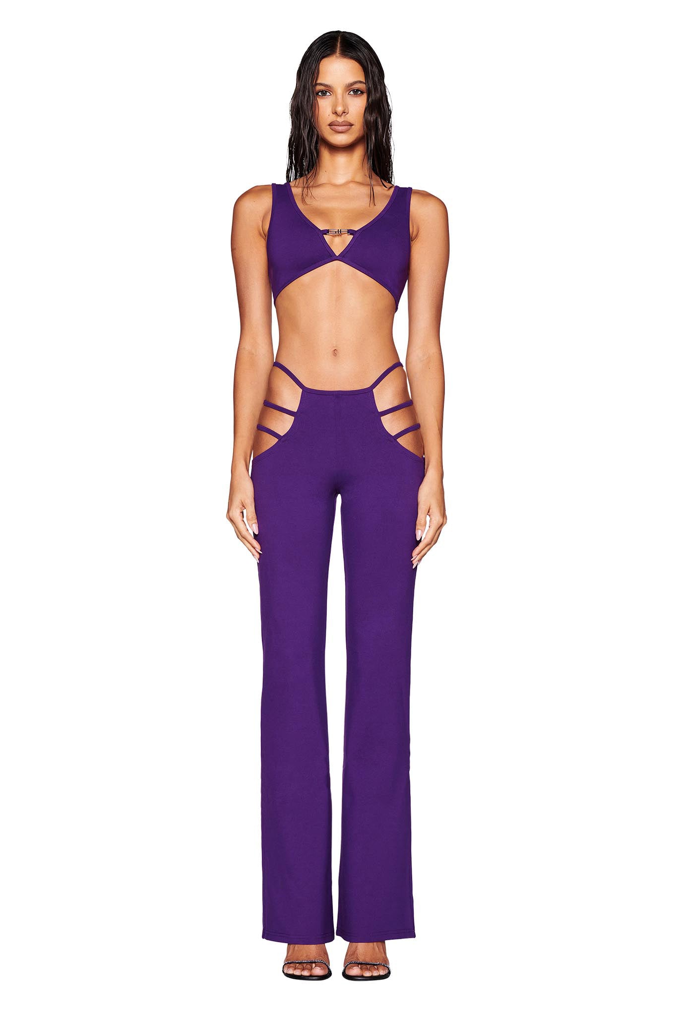 A woman poses in a body-hugging purple ensemble featuring the LUCID TOP by I.AM.GIA, which includes stylish cutouts and flared pants. The sleeveless top showcases a deep neckline, while the pants are accented with side cutouts at the hips. She finishes off her look with high-heeled sandals and slicked-back hair against a plain white backdrop.