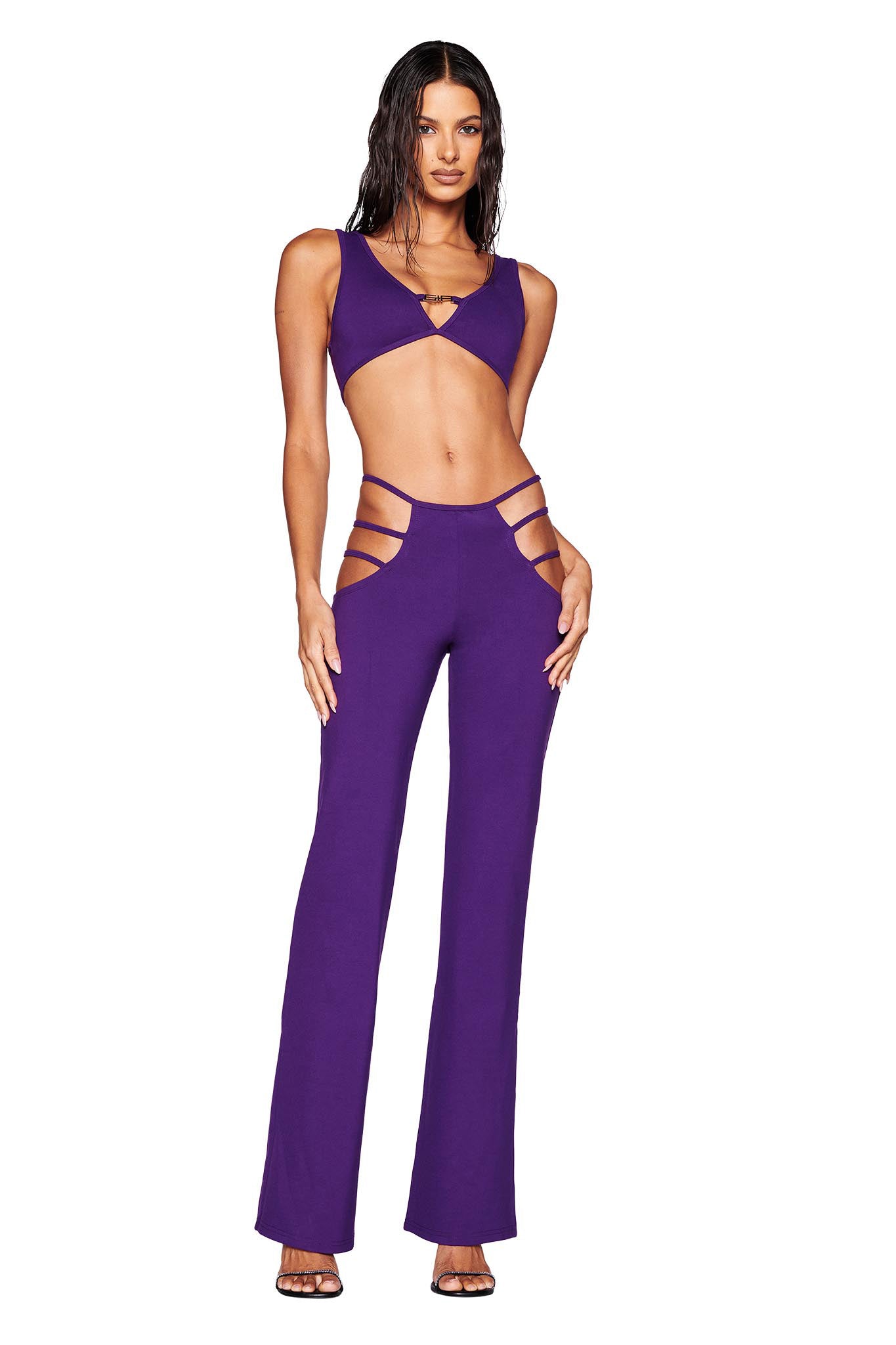 A woman poses in a striking two-piece ensemble consisting of the I.AM.GIA LUCID TOP in purple and high-waisted pants with side cutouts. Her bodycon outfit is accentuated by black heels and long, wavy hair, set against a plain white background.