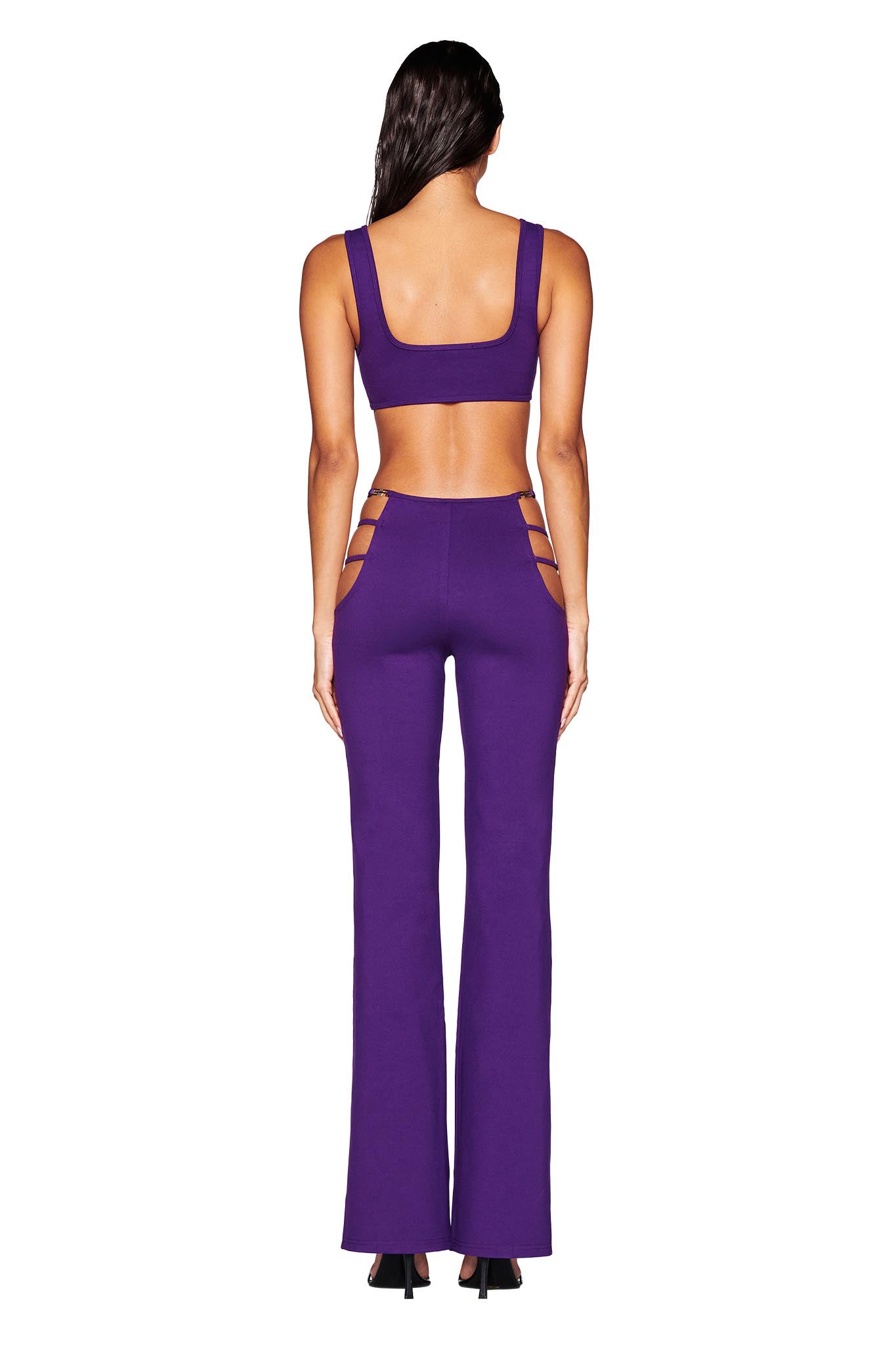 A person with long dark hair is wearing the LUCID PANT - PURPLE from I.AM.GIA, a stylish two-piece outfit with a crop top and high-rise pants featuring side hip cut-outs and an elastic waistband for added comfort as they stand facing away from the camera.