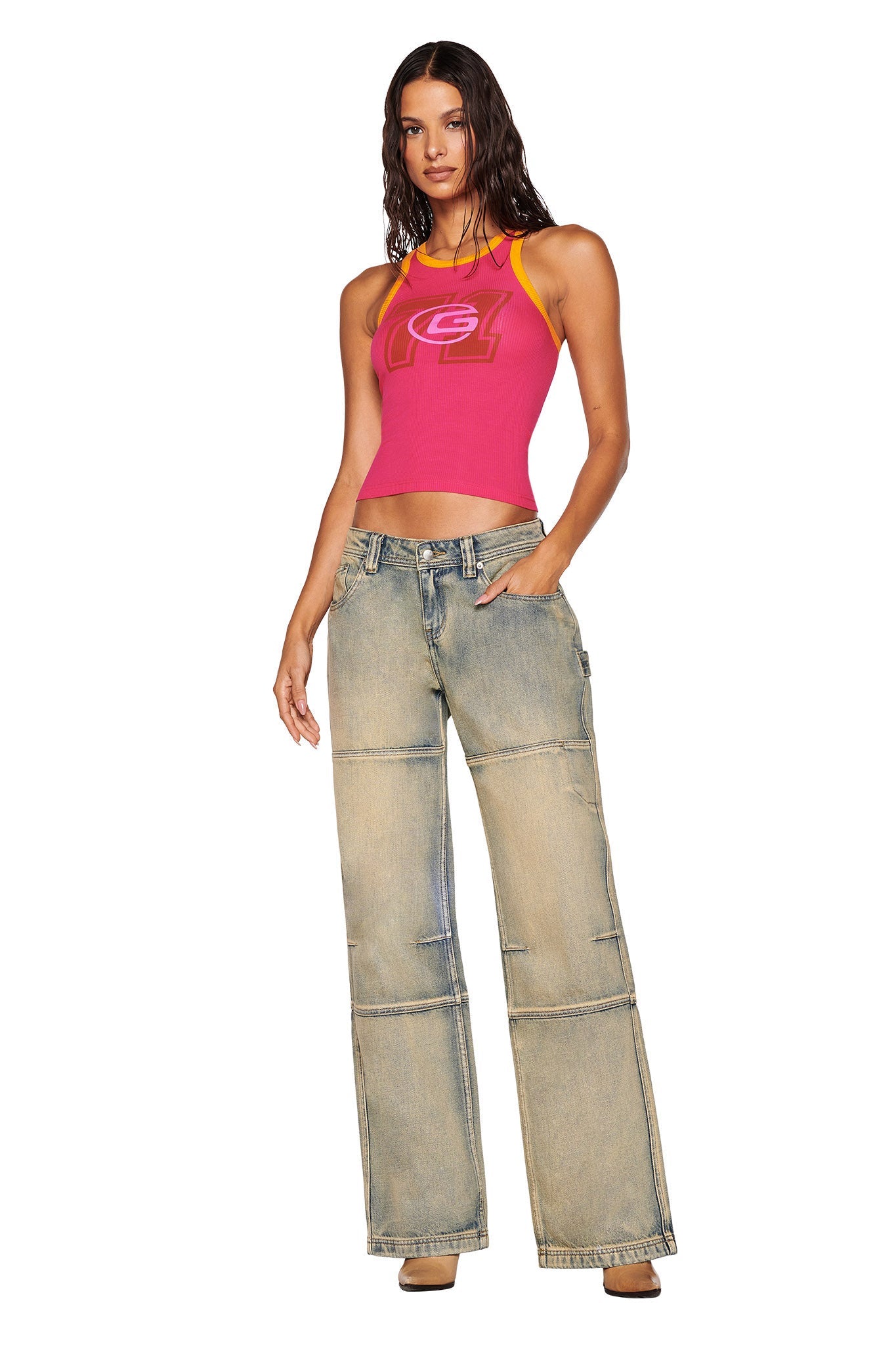 A person stands confidently in a hot pink I.AM.GIA PENELOPE TANK - PINK, paired with light blue wide-legged jeans. One hand rests casually in their pocket, and their long, wavy hair cascades against the plain white background.