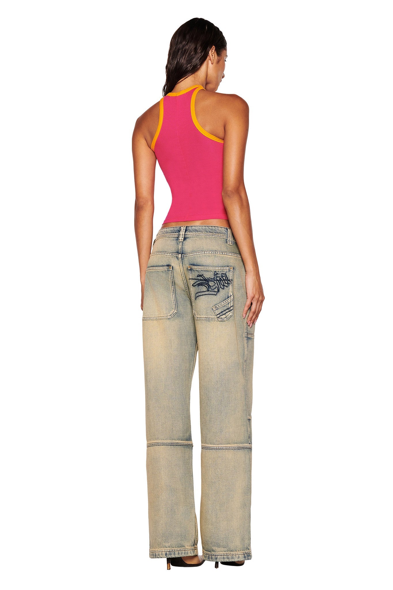 A woman stands with her back to us, wearing an I.AM.GIA PENELOPE TANK in hot pink and wide-legged, light-wash jeans featuring distressed details and patches on the back pockets. Her dark hair is worn loose, enhancing the contrast with her dark-colored shoes against a simple white background.