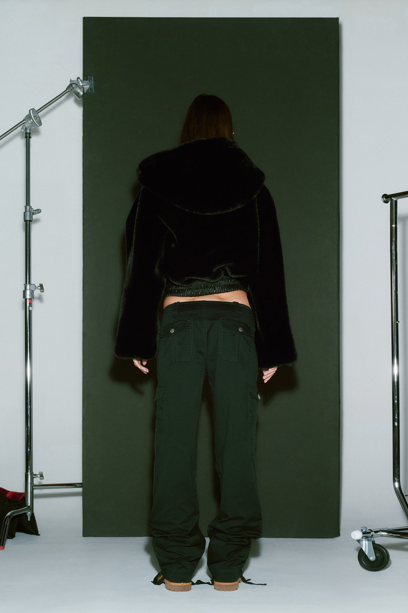 A person stands with their back to the camera, wearing an I.AM.GIA 717 JACKET - BLACK paired with dark pants. The setting appears to be a studio, featuring equipment such as a C-stand and a wheeled clothing rack visible on either side of a black backdrop.