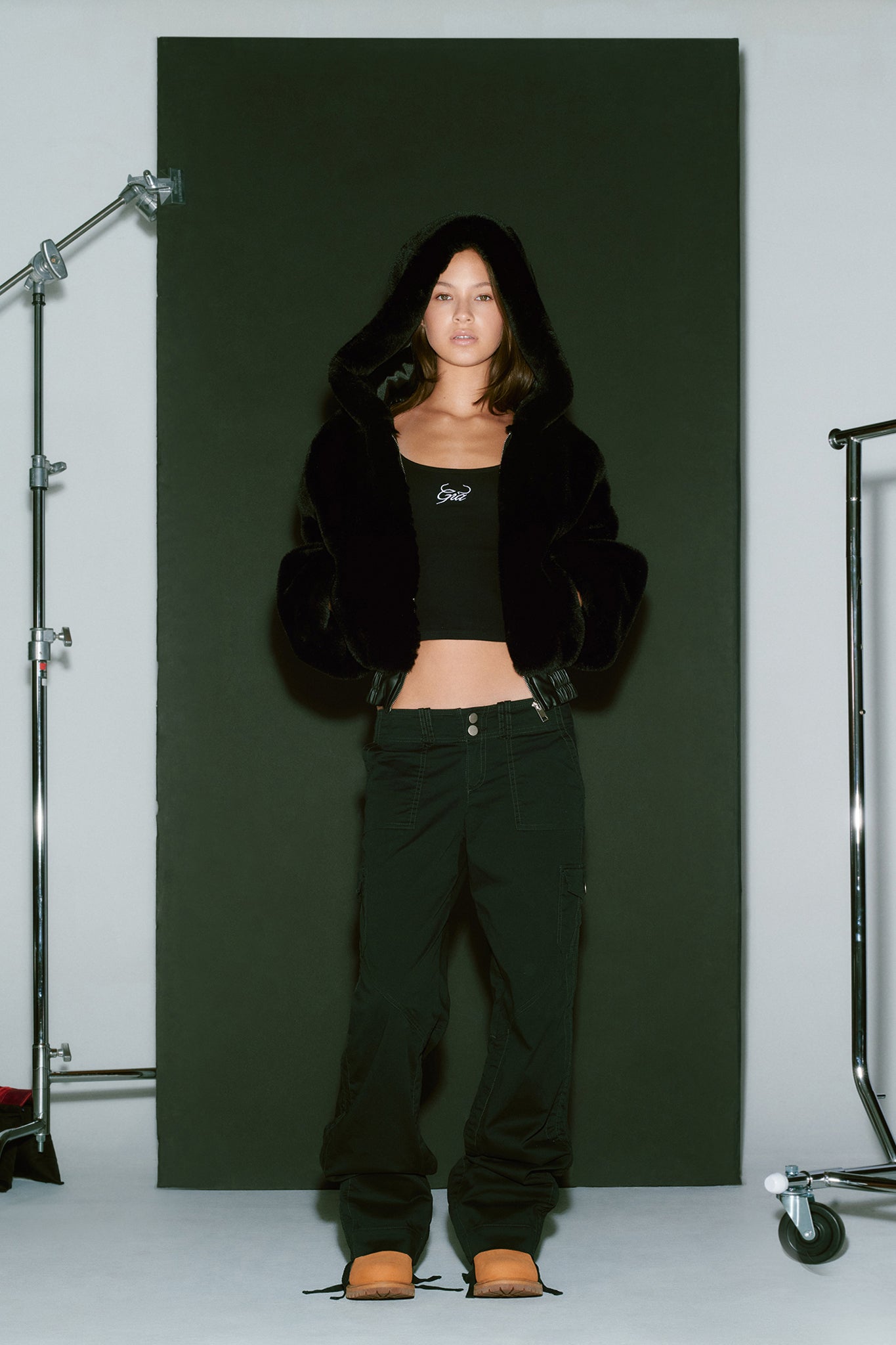 A young woman, dressed head-to-toe in I.AM.GIA apparel, stands against a black background. She's wearing the 717 JACKET - BLACK with the hood up, a black crop top, and black pants. With her hands in her pockets, she gazes directly at the camera. Studio lighting equipment is partially visible on the sides.