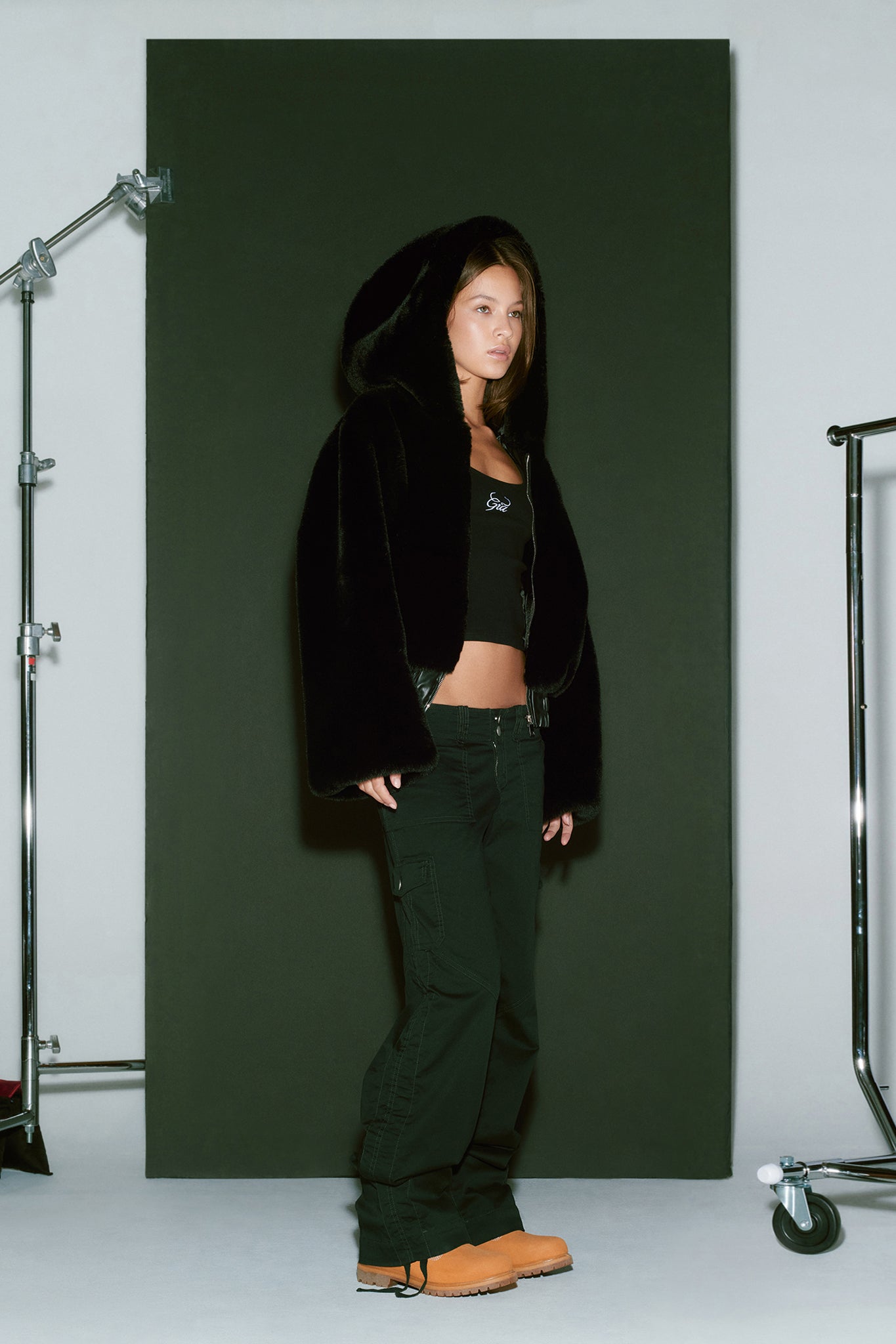 A woman stands in front of a dark backdrop, dressed head-to-toe in I.AM.GIA. She wears the 717 JACKET - BLACK, along with a black crop top and black pants, paired with tan boots. Studio equipment, including light stands and a hanger rack, is visible on either side of her.