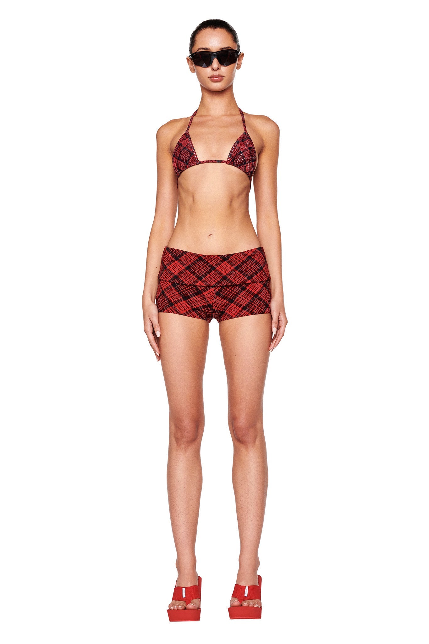 A woman stands facing forward, wearing a red plaid bikini top and matching high-waisted plaid shorts from I.AM.GIA. She also has on SHELBY FLIP FLOP - RED from I.AM.GIA and dark sunglasses. The background is plain white.