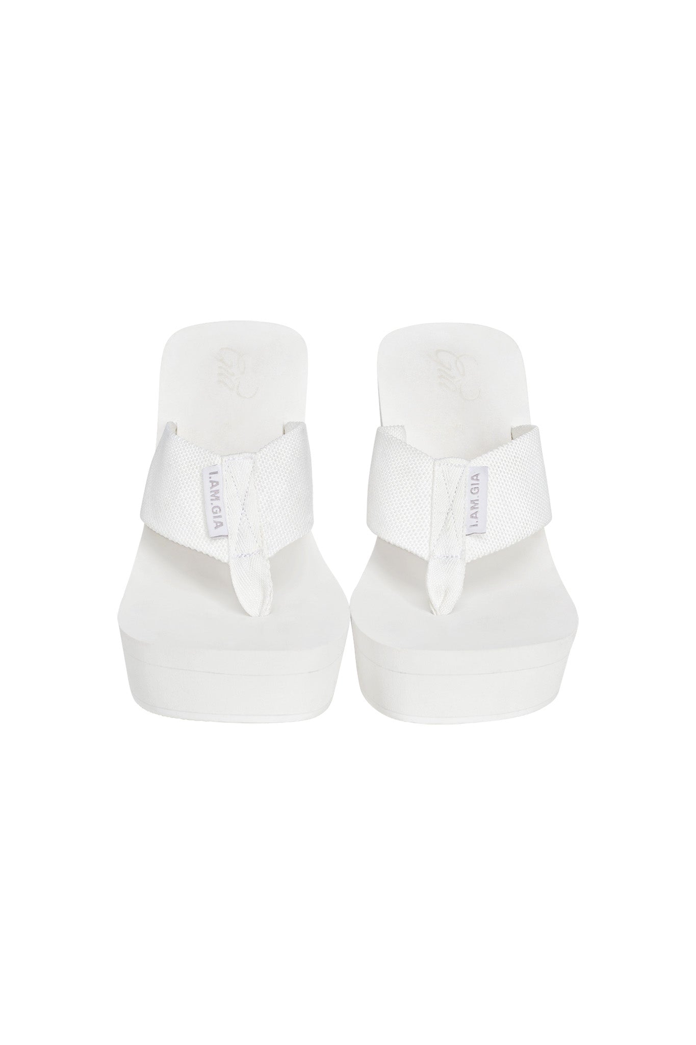 A pair of white SHELBY FLIP FLOPS from I.AM.GIA with a thick sole and wide straps features the brand name "MADEGA" printed on the straps in purple. These stylish flip-flops, embodying I.AM.GIA's bold aesthetic, are positioned side by side against a plain white background.