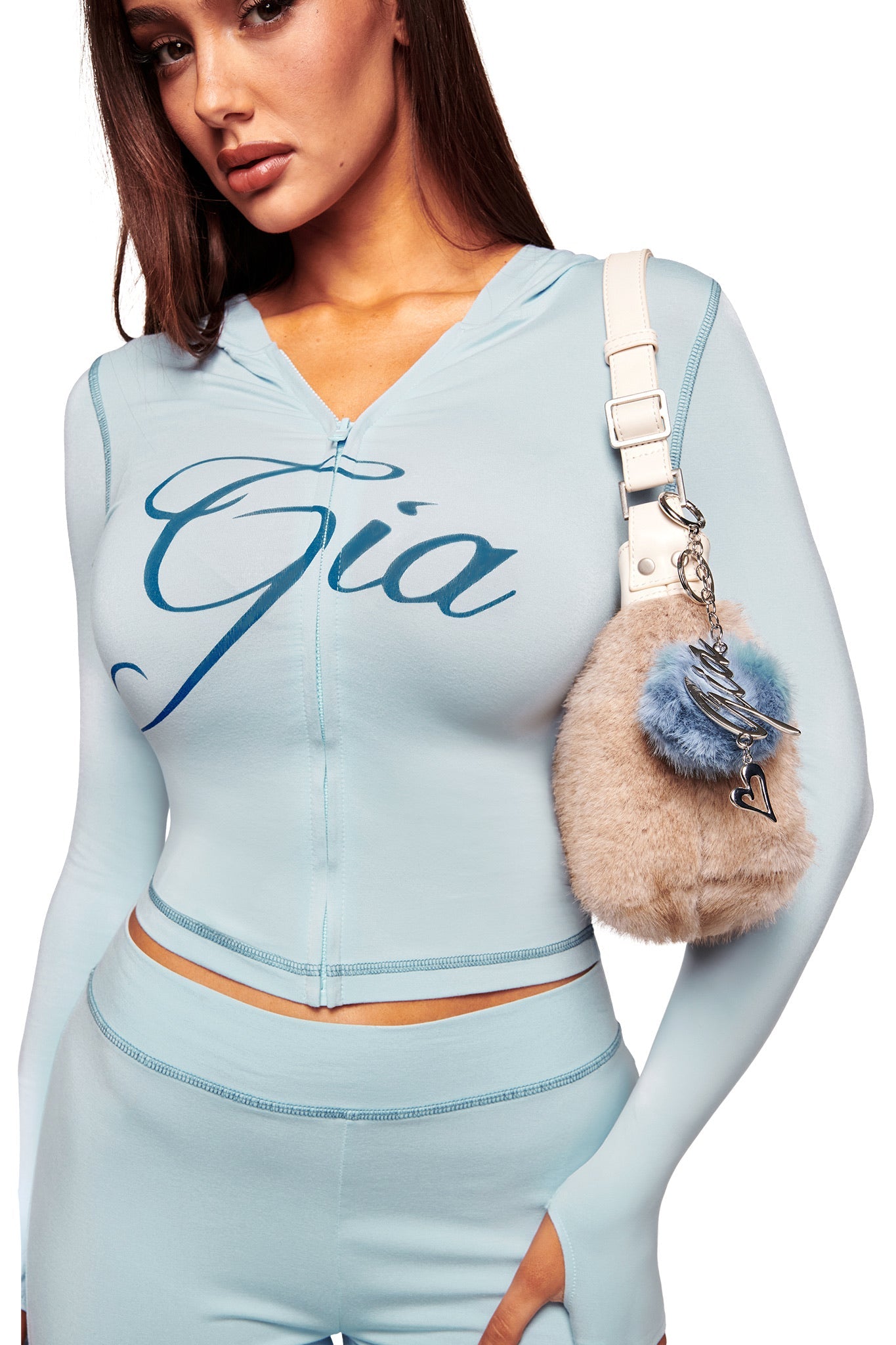 A woman in a light blue I.AM.GIA outfit carries a beige and blue furry bag adorned with the LARSA KEYCHAIN - BLUE and a heart charm, posing stylishly against a white background.