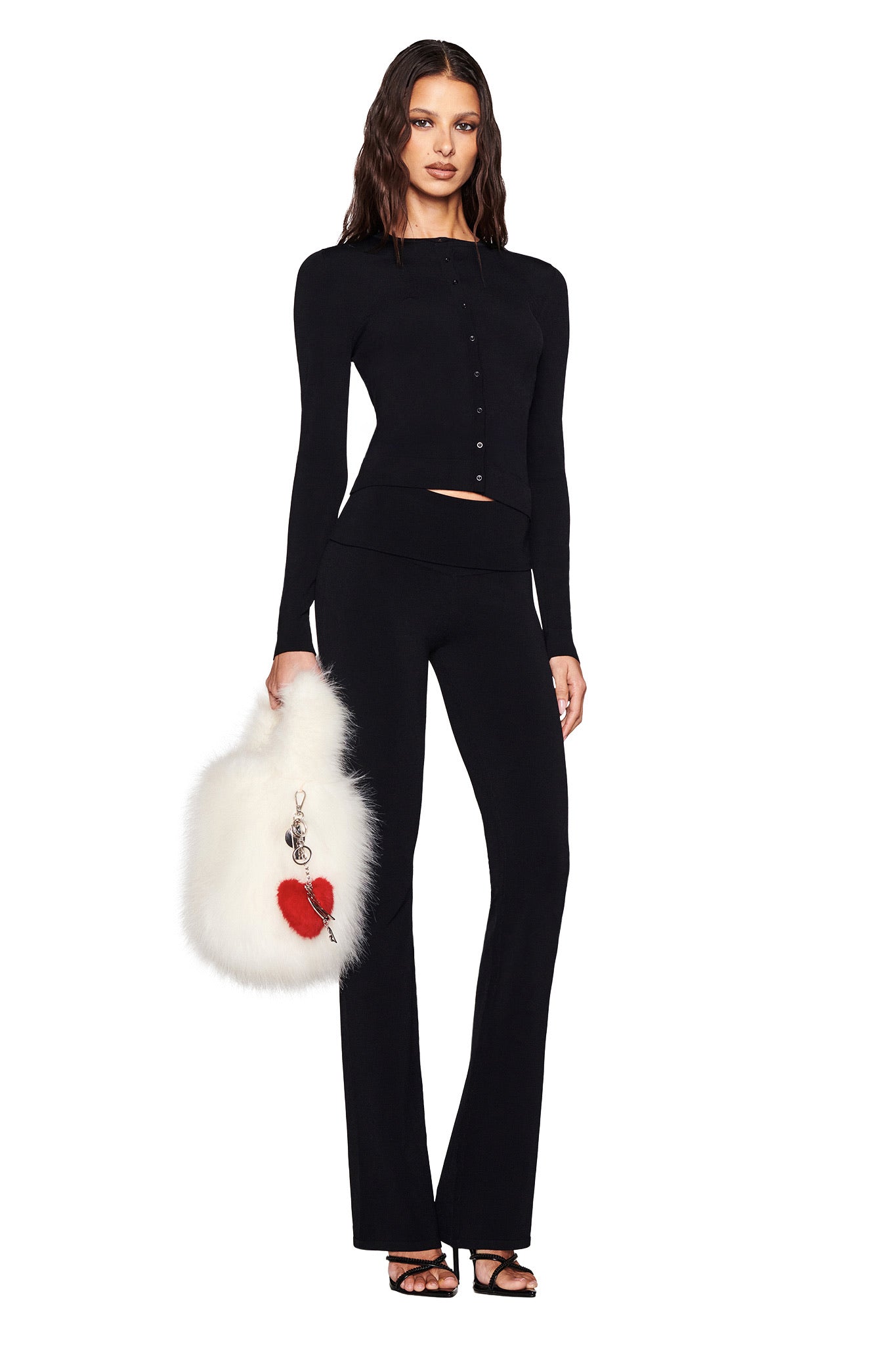 A person in a fitted black outfit with a button-up top and trousers holds a white faux fur bag embellished with red hearts. With long dark hair and black open-toe heels, they showcase the I.AM.GIA VALENTINA KEYCHAIN - RED on the bag, against a simple white background.