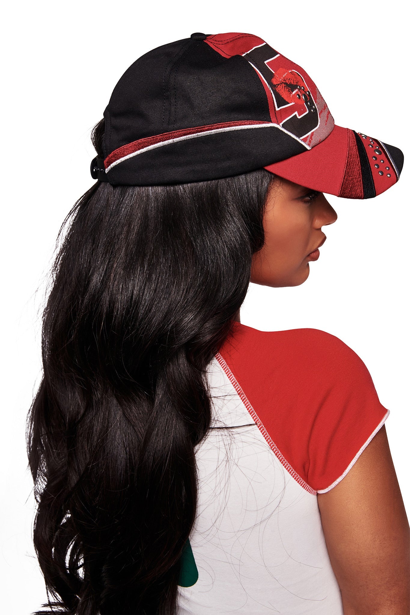 A woman with long, wavy black hair faces away in front of a white background, wearing the I.AM.GIA SERENA HAT - RED and a matching short-sleeve shirt with red, black, and white panels.
