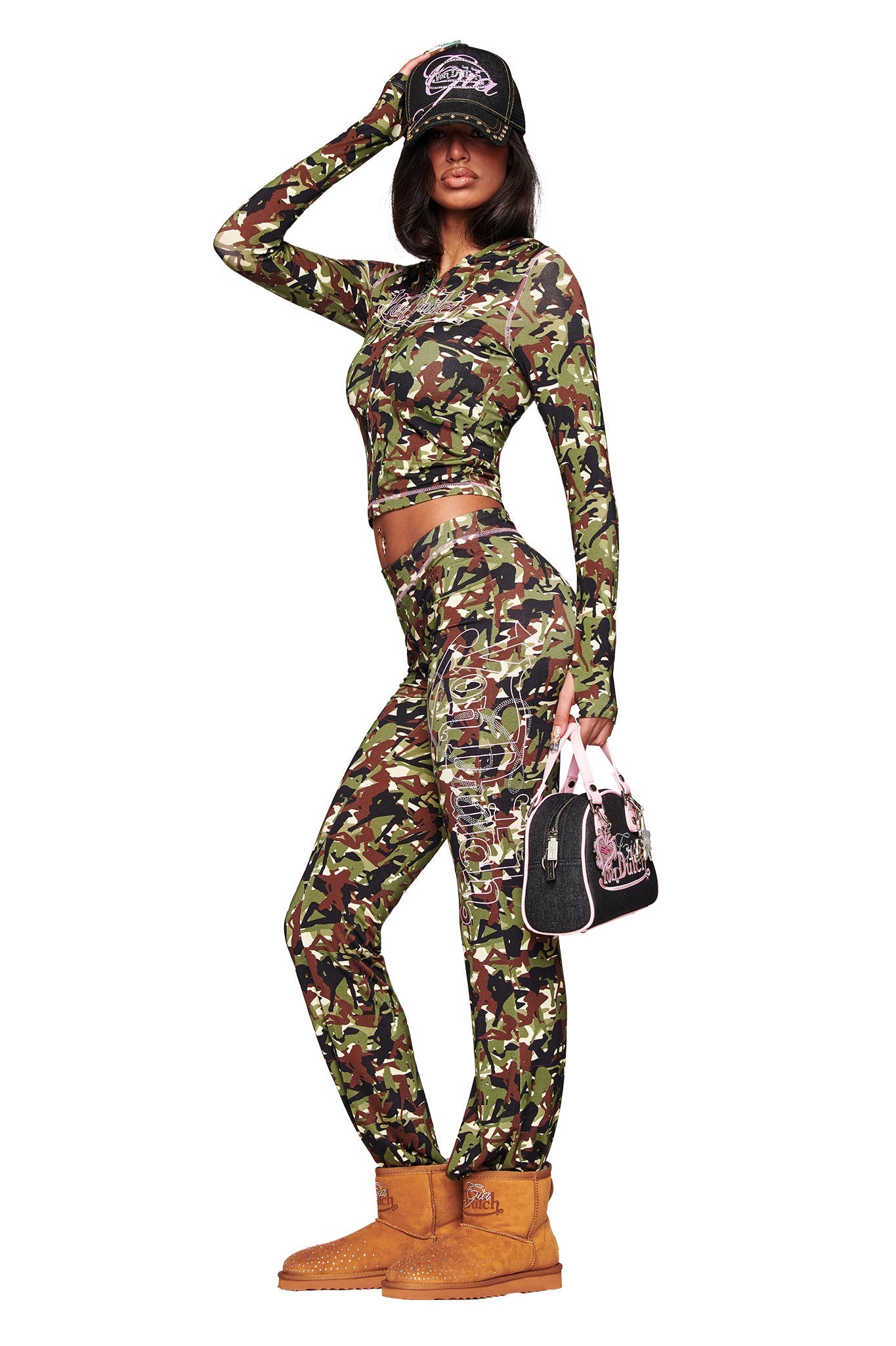 A woman poses in the I.AM.GIA X VON DUTCH BLARE HOODIE - GIA GIRL CAMO with matching pants, tan boots, and a black cap. She holds a small black Von Dutch handbag with pink accents against a white background.