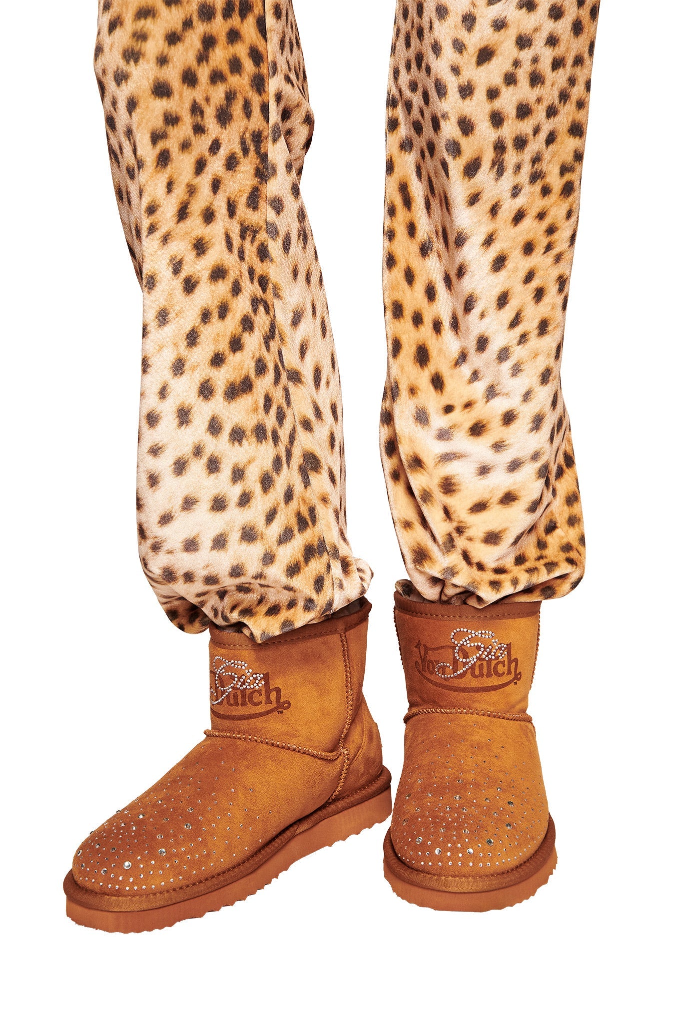 A person wears leopard-print pants and brown I.AM.GIA X VON DUTCH Faux Shearling Boots, featuring rhinestone studs and embroidered cuffs.