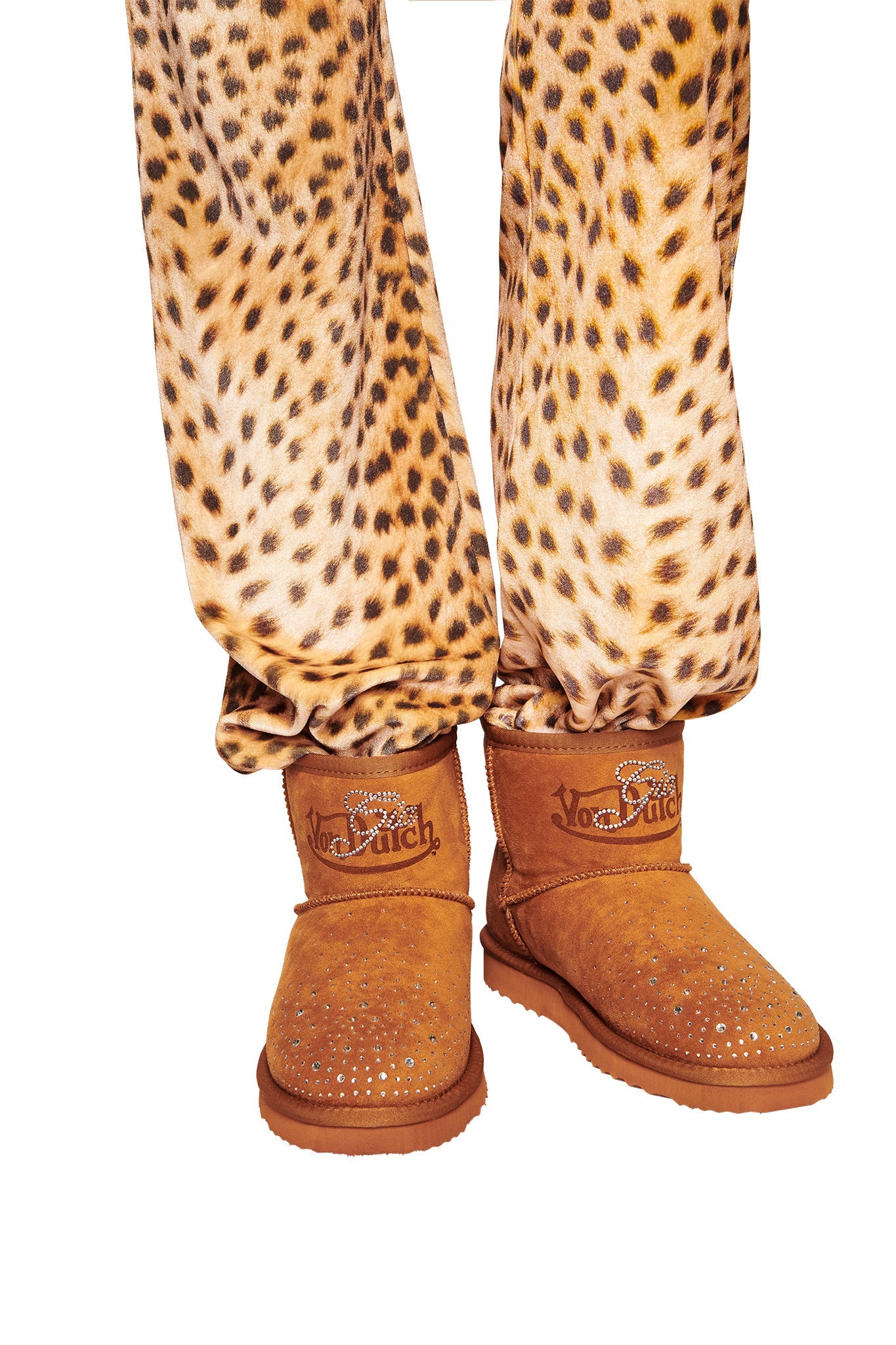 A person wears leopard print pants and I.AM.GIA X VON DUTCH Faux Shearling Boots in Brown. The photo is cropped to show only the lower legs and feet against a white background.