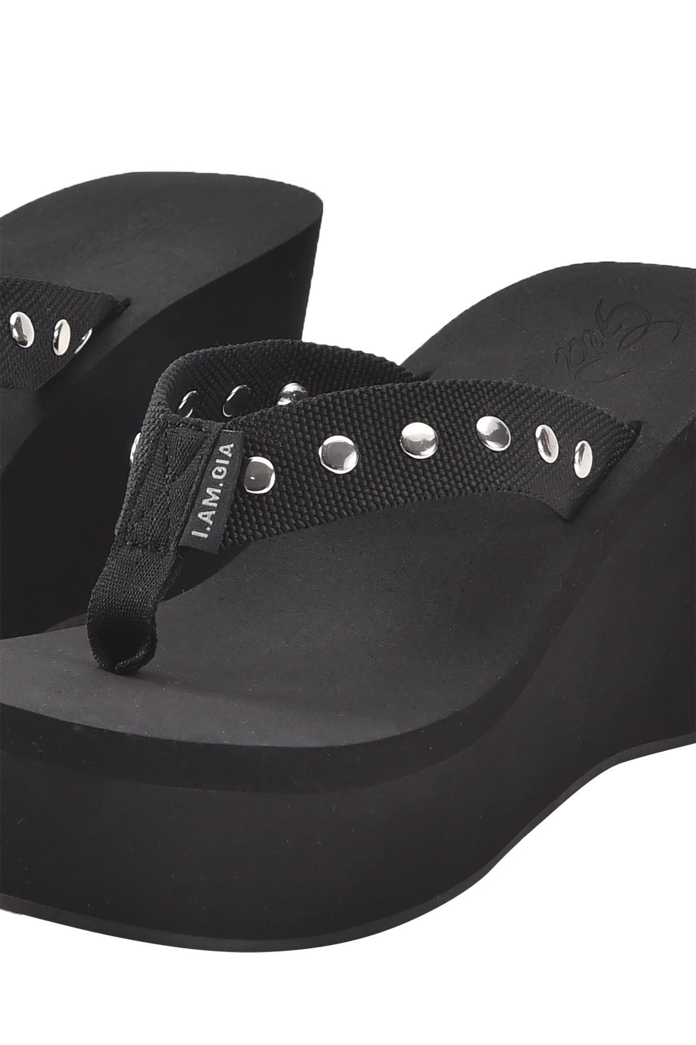 A close-up of the SHELBY STUDDED FLIP FLOP 2.0 - BLACK by I.AM.GIA, a black wedge sandal with fabric straps, metal eyelets, and a thick platform sole for extra height and style.