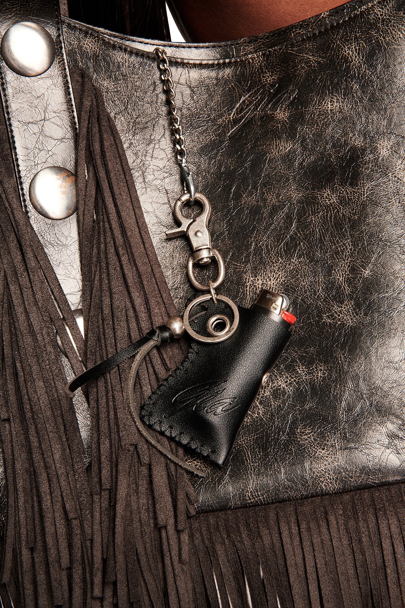 Close-up of a metallic textured leather bag with fringe and silver buttons, featuring the I.AM.GIA VENUS LIGHTER KEYCHAIN - BLACK on a chain. A subtle GIA logo accents the stylish design.