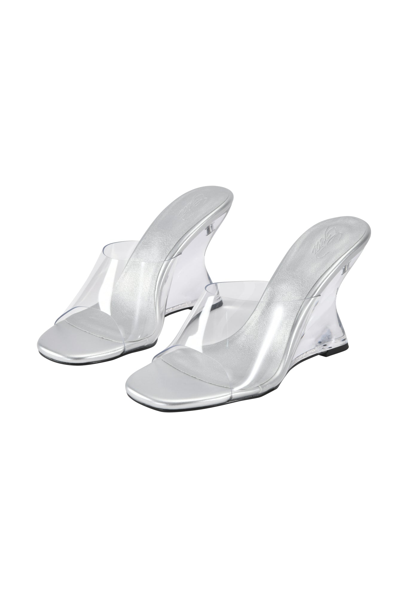 The I.AM.GIA RHEA CLEAR WEDGE HEEL – TRANSPARENT features high-heeled mule sandals with clear vinyl straps, silver metallic insoles, a transparent wedge heel, and open toes, showcased against a white background.