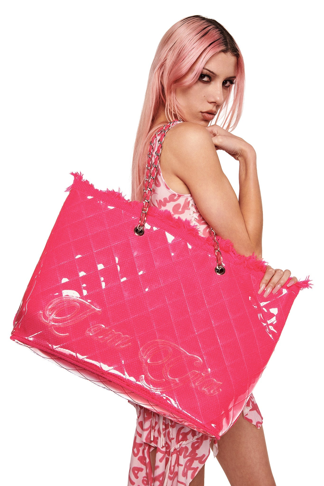 A person with long pink hair carries the I.AM.GIA DANNI TOTE BAG in hot pink, featuring chain straps, over their shoulder while wearing a pink and white patterned outfit against a white background.