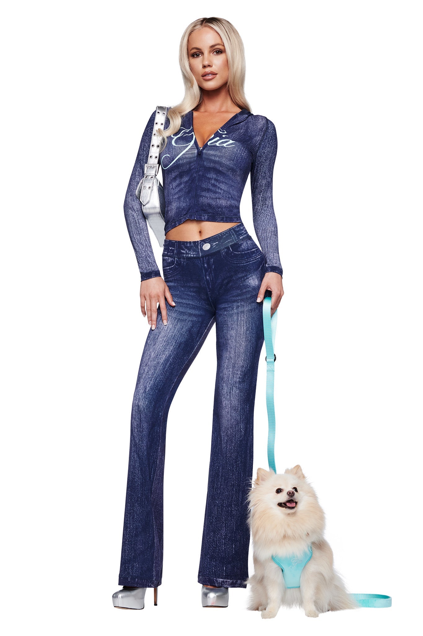 A woman in a fitted I.AM.GIA blue denim-style top and flared pants, silver heels, and holding a silver purse walks her small white dog, who wears the STAR PAWFECT 3-PIECE SET - BABY BLUE with matching harness and leash.