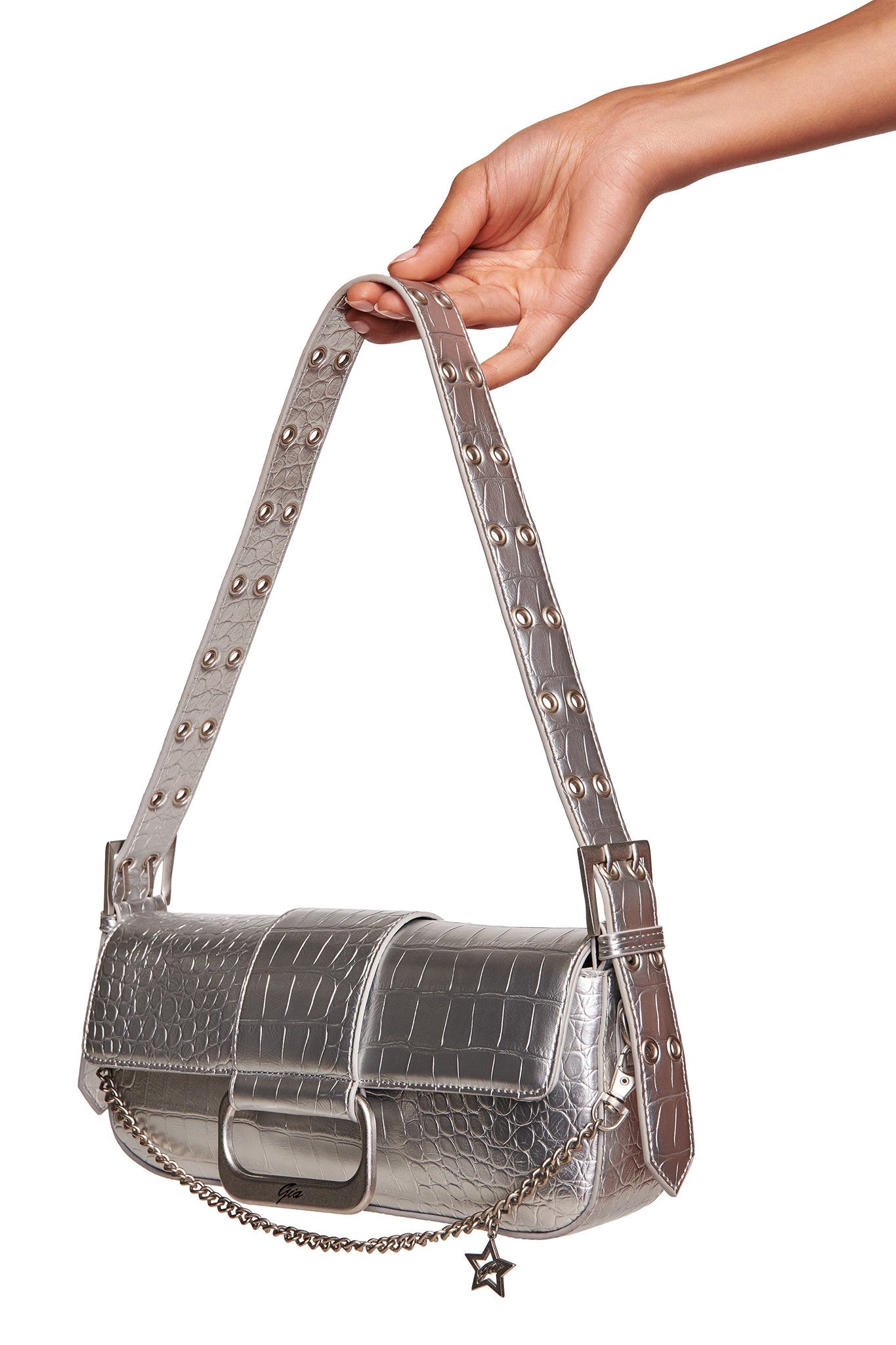 A hand holds the I.AM.GIA MILA METALLIC BAG in silver with faux crocodile texture, studded strap, front flap closure, and a chain featuring a star charm.