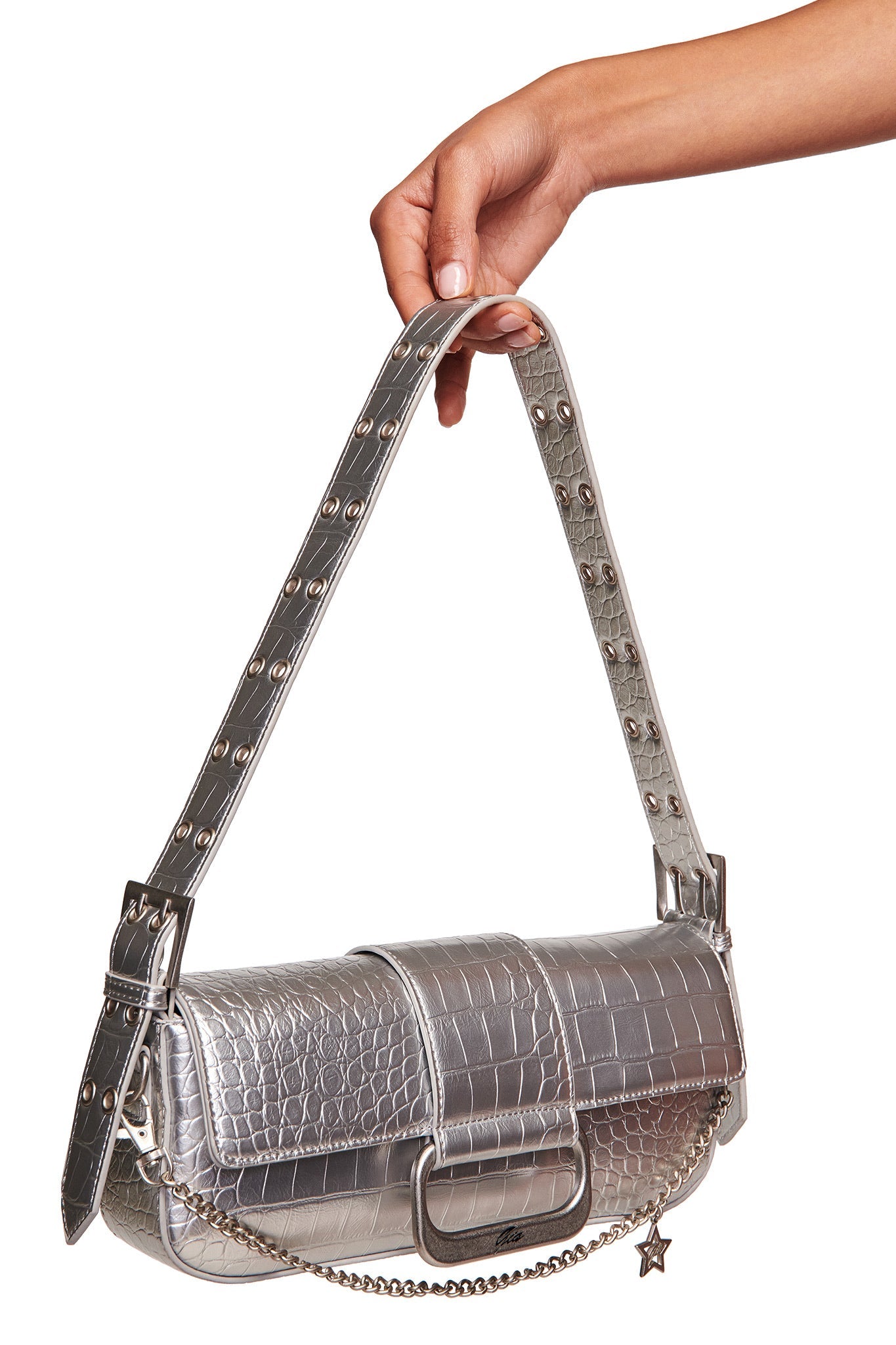A hand holds the MILA METALLIC BAG - SILVER METALLIC by I.AM.GIA, featuring faux crocodile texture, a wide studded strap, chain detail, and star charm against a white background.
