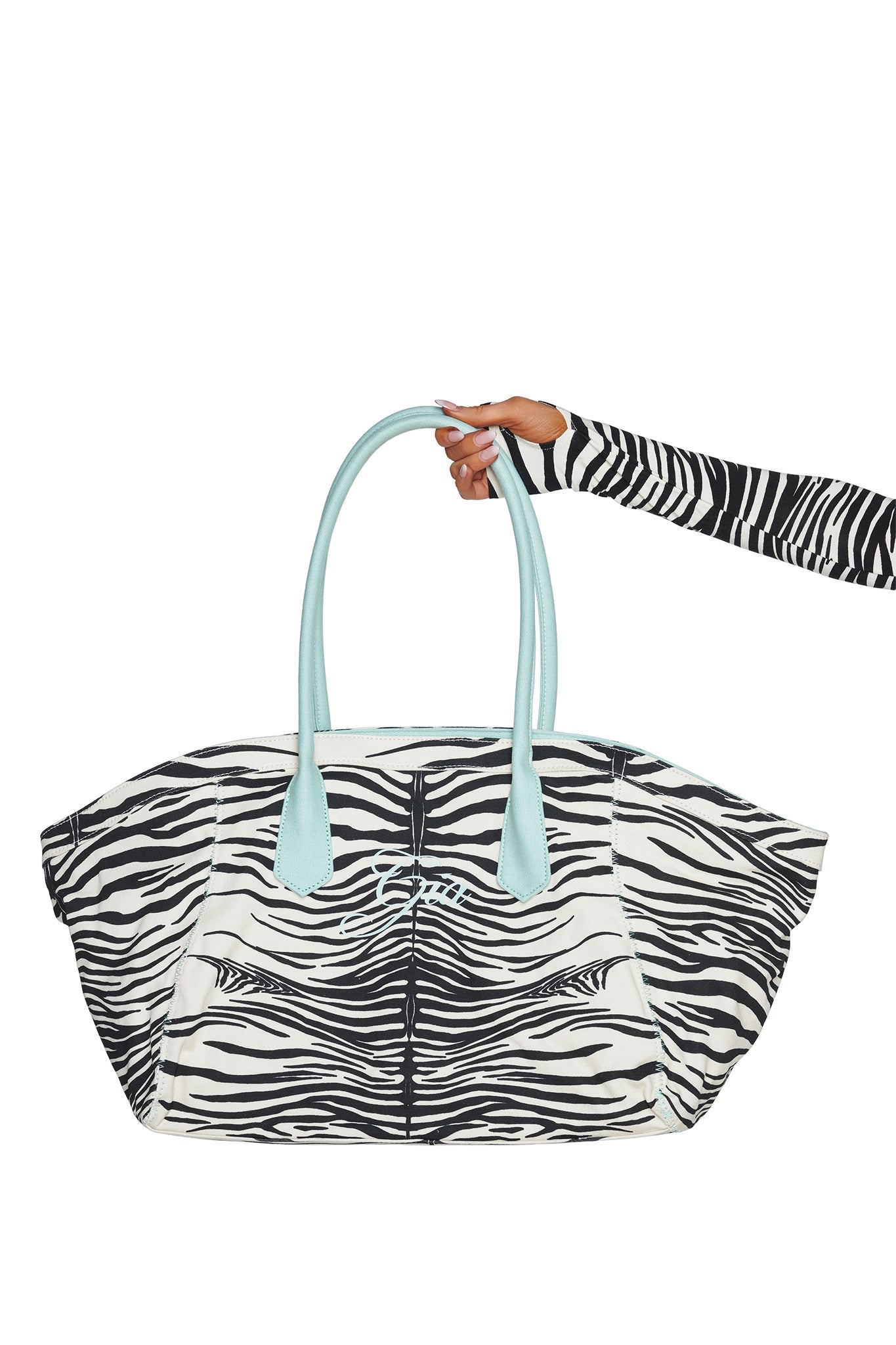 A hand in a zebra-striped sleeve holds the I.AM.GIA BLARE TOTE BAG - FINE LINE ZEBRA, an oversized tote crafted from heavyweight cotton canvas with light blue handles and a matching light blue logo on the front, set against a white background.