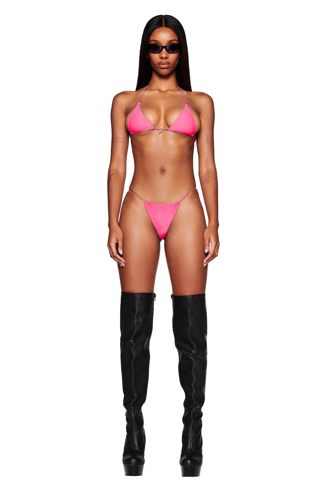 A person is standing against a white background, dressed in I.AM.GIA's BAMBI BIKINI BOTTOM in hot pink paired with black knee-high boots. They have long, straight hair and are wearing dark sunglasses, with their arms resting by their sides.