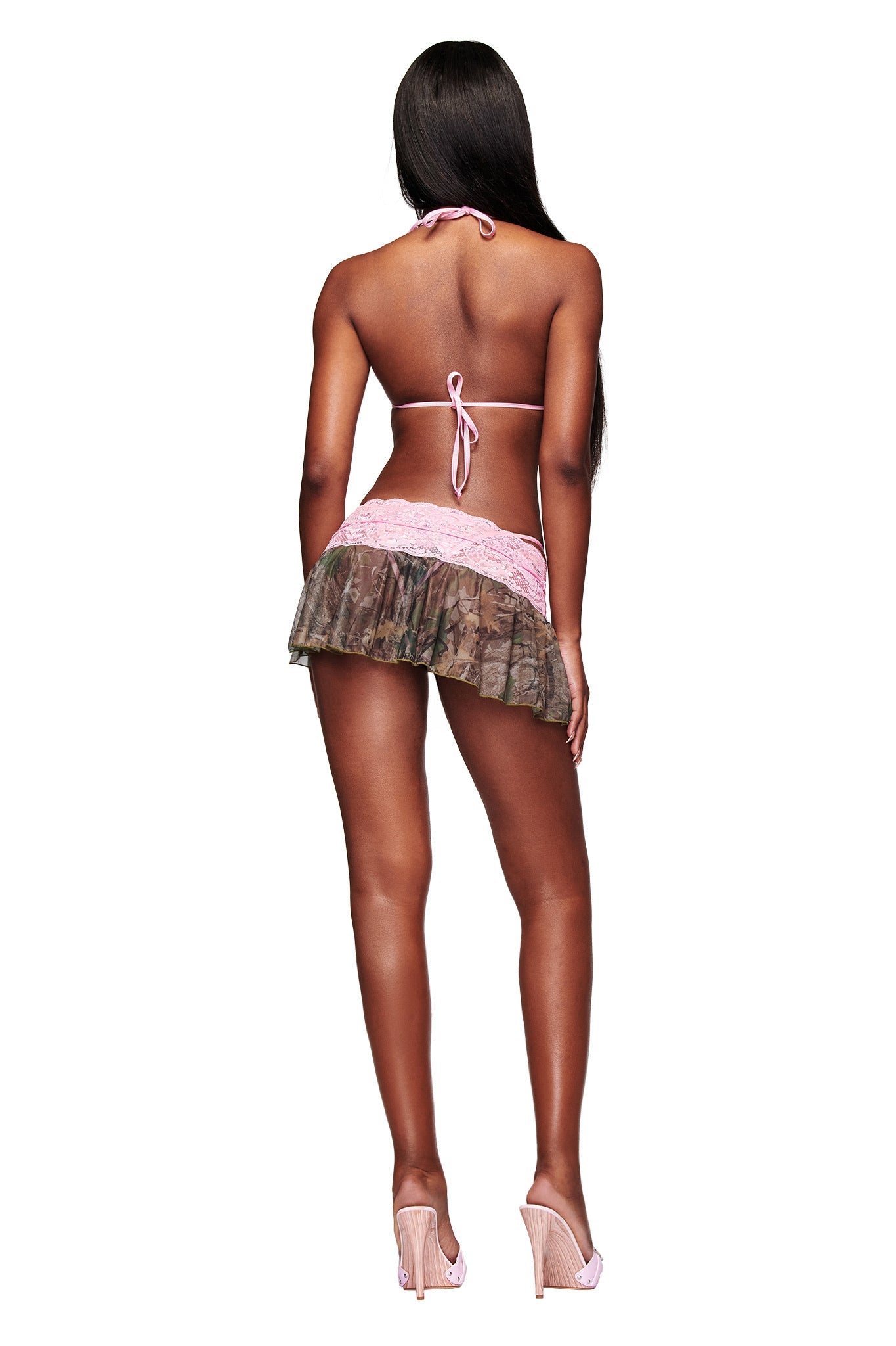 A woman with long, straight hair and high heels poses facing away in the I.AM.GIA SAKINA BIKINI TOP - BARK AND BITE CAMO and a short camo mini skirt with sequin lace trim against a white background.