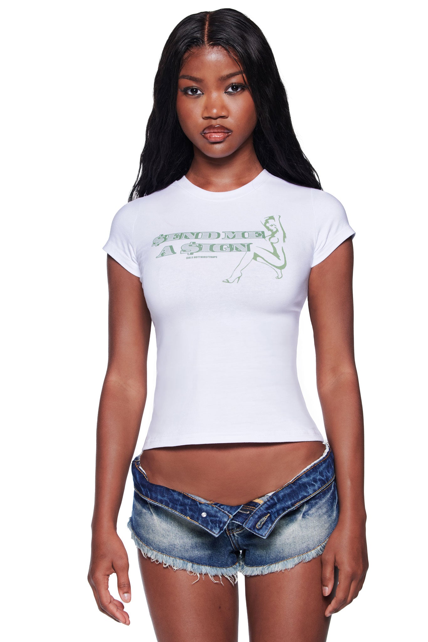 A woman with long dark hair wears the I.AM.GIA POLINA DOLLA SIGN 2.0 TEE in white and low-rise, partially unbuttoned denim shorts, standing against a plain white background.