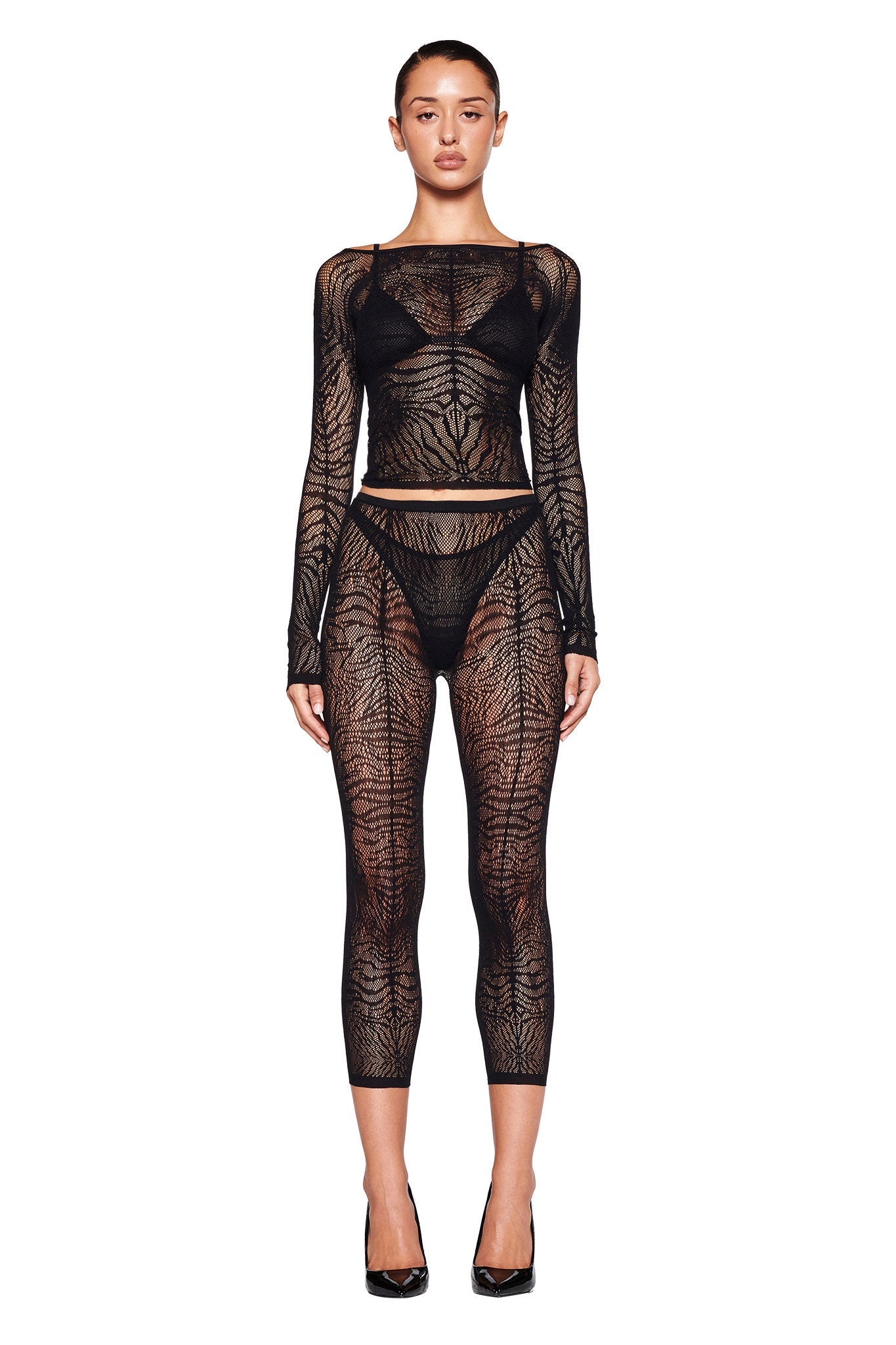 A woman stands facing the camera, wearing the I.AM.GIA SYBIL CAPRI - BLACK : ZEBRA bodysuit. The sheer, black bodycon silhouette features a high neckline with an intricate lace-like design, revealing undergarments underneath. She is paired with black high-heeled shoes against a white background.