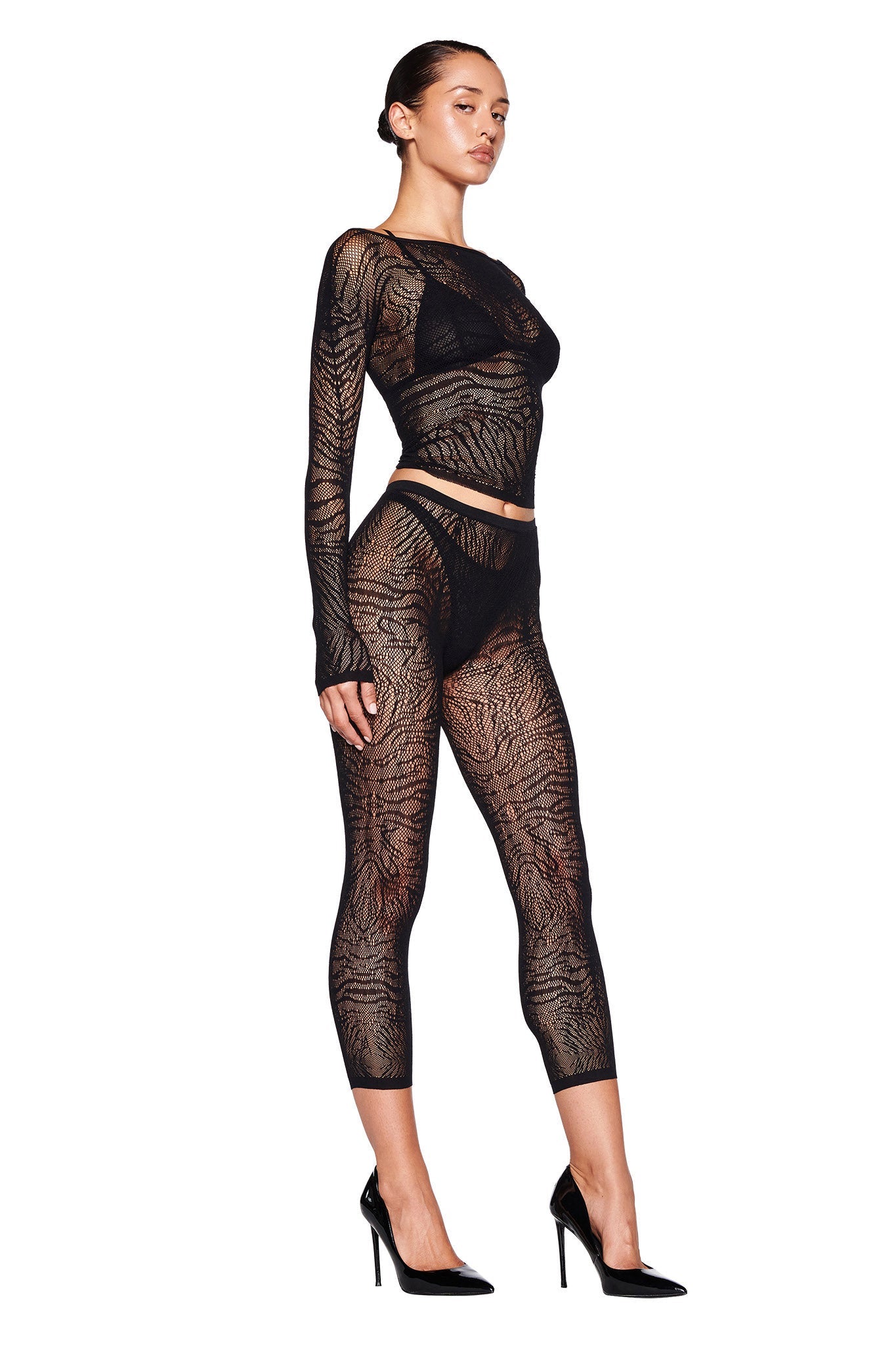 A person poses in the SYBIL CAPRI - BLACK : ZEBRA jumpsuit by I.AM.GIA, featuring intricate zebra-like patterns. The sheer black outfit has long sleeves and ends just above the ankle. The person complements the look with black high-heeled shoes and has their hair tied back. The background is plain white.