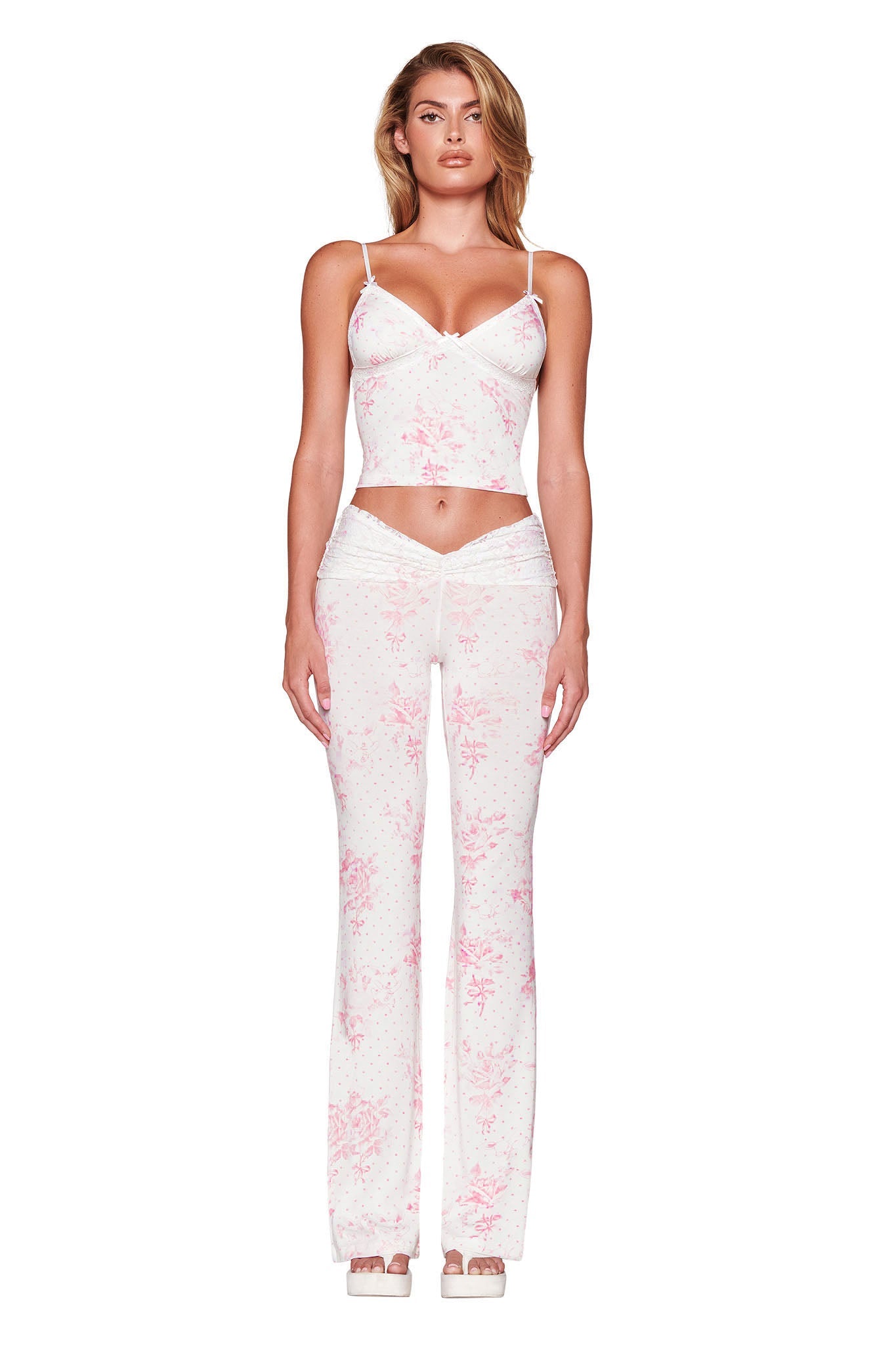 A woman is standing against a white background wearing the I.AM.GIA KENDRA PANT - WHITE : FLORAL: BUNNY BOUQUET ensemble, featuring a custom floral print crop top with spaghetti straps and matching high-waisted pants. Her hands rest at her sides as her wavy hair cascades over her shoulders. She completes the look with stylish high-heeled sandals.