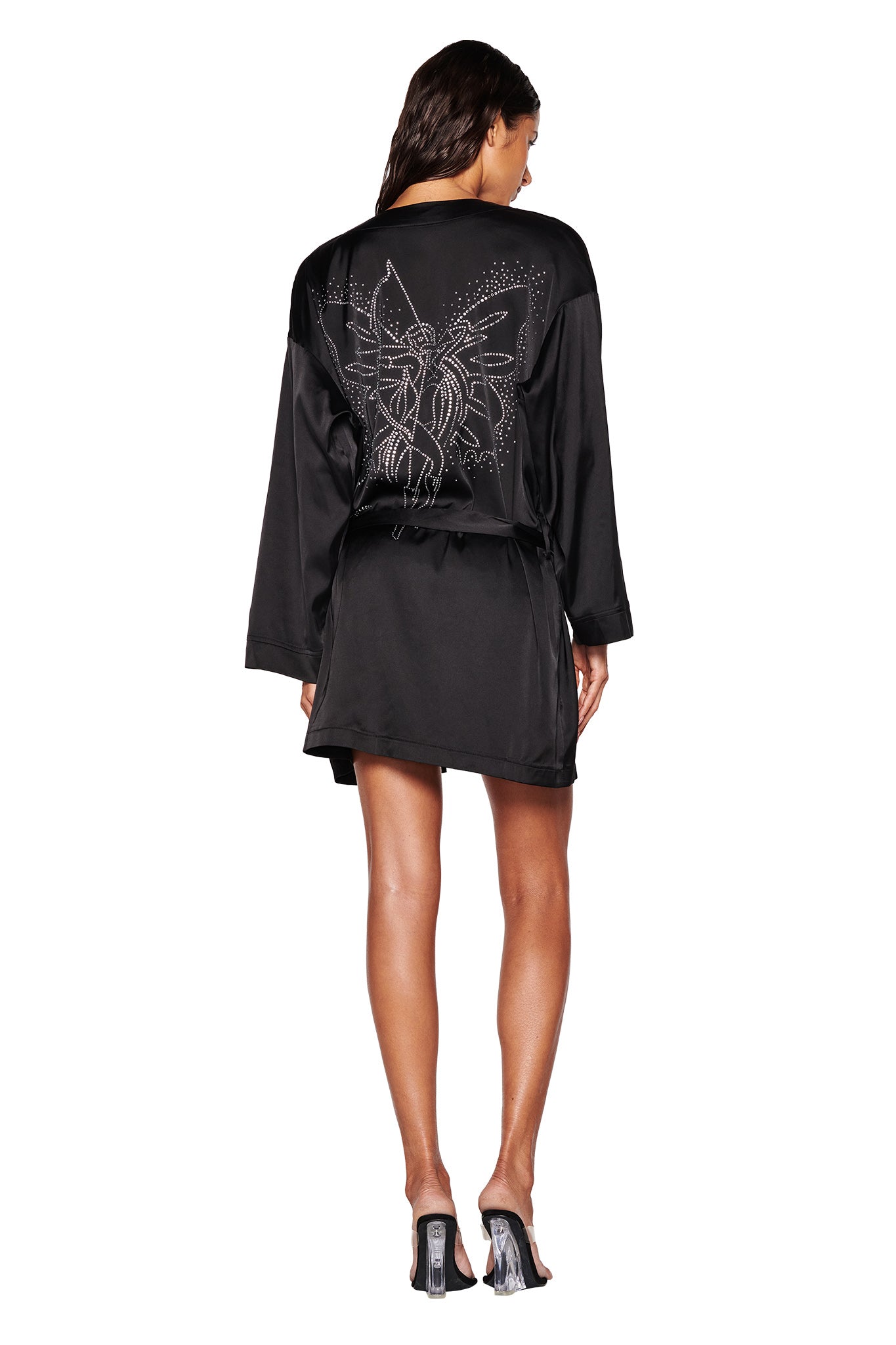 A person faces away from the camera, wearing the I.AM.GIA LARSA 2.0 Sleep Robe in black. The satin fabric is adorned with an elaborate silver embroidery that resembles butterfly diamante artwork. Their long hair flows down as they wear a pair of clear-heeled shoes.