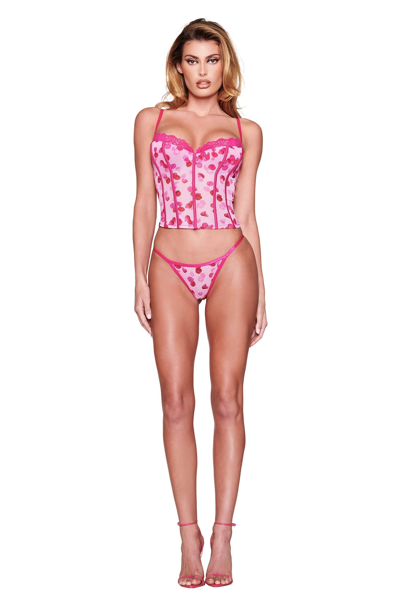 A person poses against a white background wearing the ROSALIA BUSTIER SET - PINK: PRETTY PETAL from I.AM.GIA, featuring heart patterns and matching bottoms, paired with pink high heels.