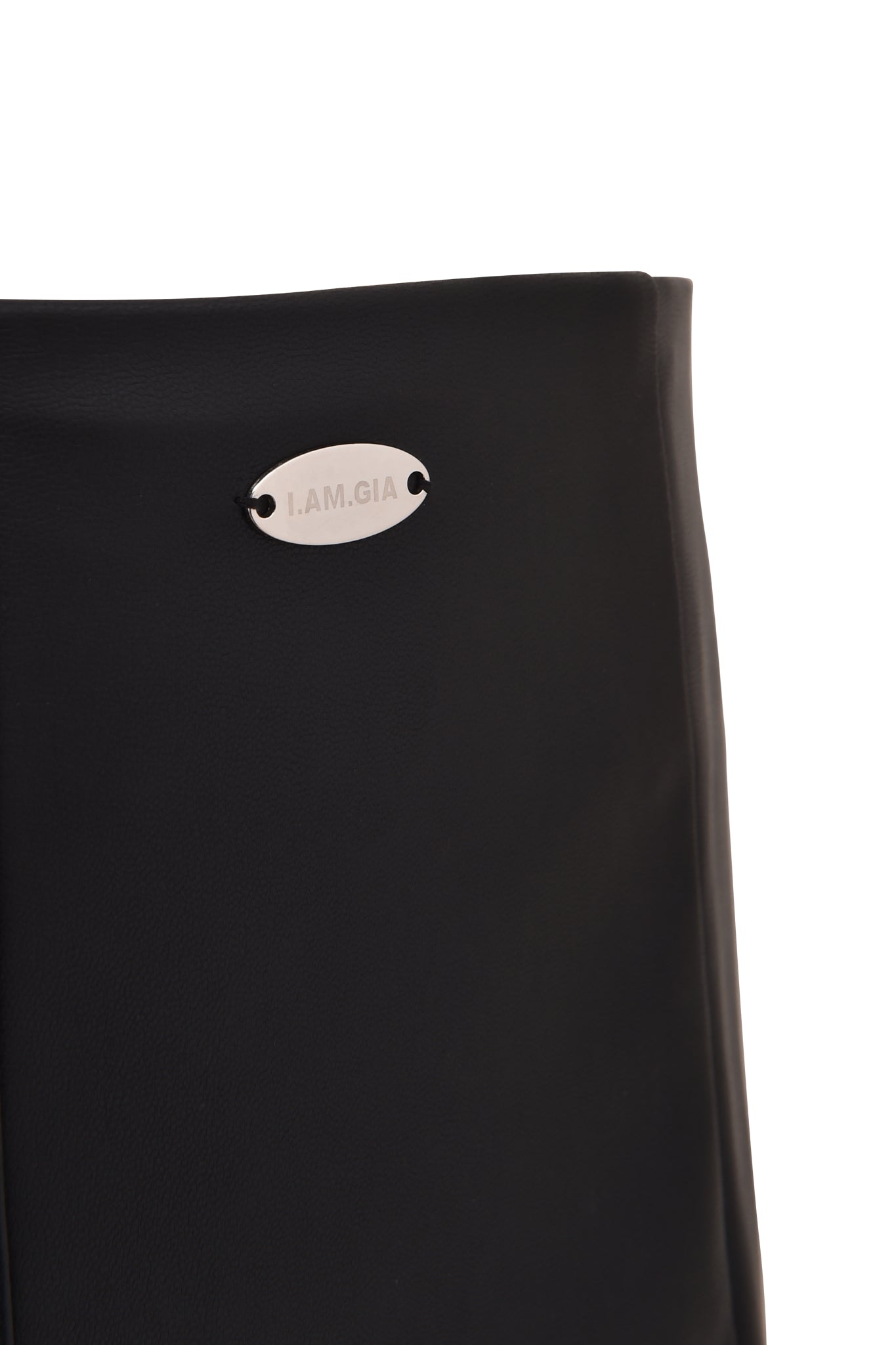 Close-up of the TEMPEST HOT SHORT - BLACK by I.AM.GIA, featuring black PU material and a small, oval silver metal tag reading "I.AM.GIA" attached near the top edge.