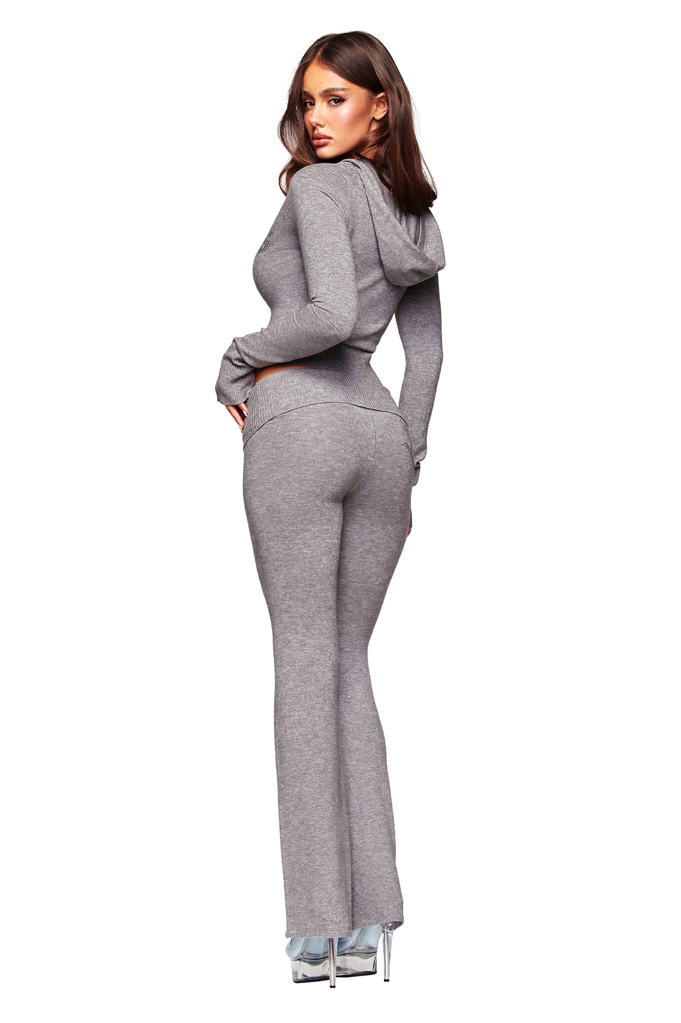 A woman faces away, glancing over her shoulder in a fitted, light gray hooded top and matching I.AM.GIA JESSA PANT with flared legs, styled with clear high heels against a white background.