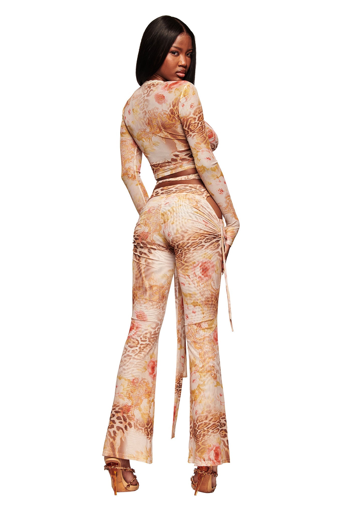 A woman poses with her back slightly turned, wearing the I.AM.GIA ERISKA TOP - DESERT ROSE, a fitted long-sleeve mesh wrap top in a floral animal print, matched with flared pants and high-heeled sandals against a white background.