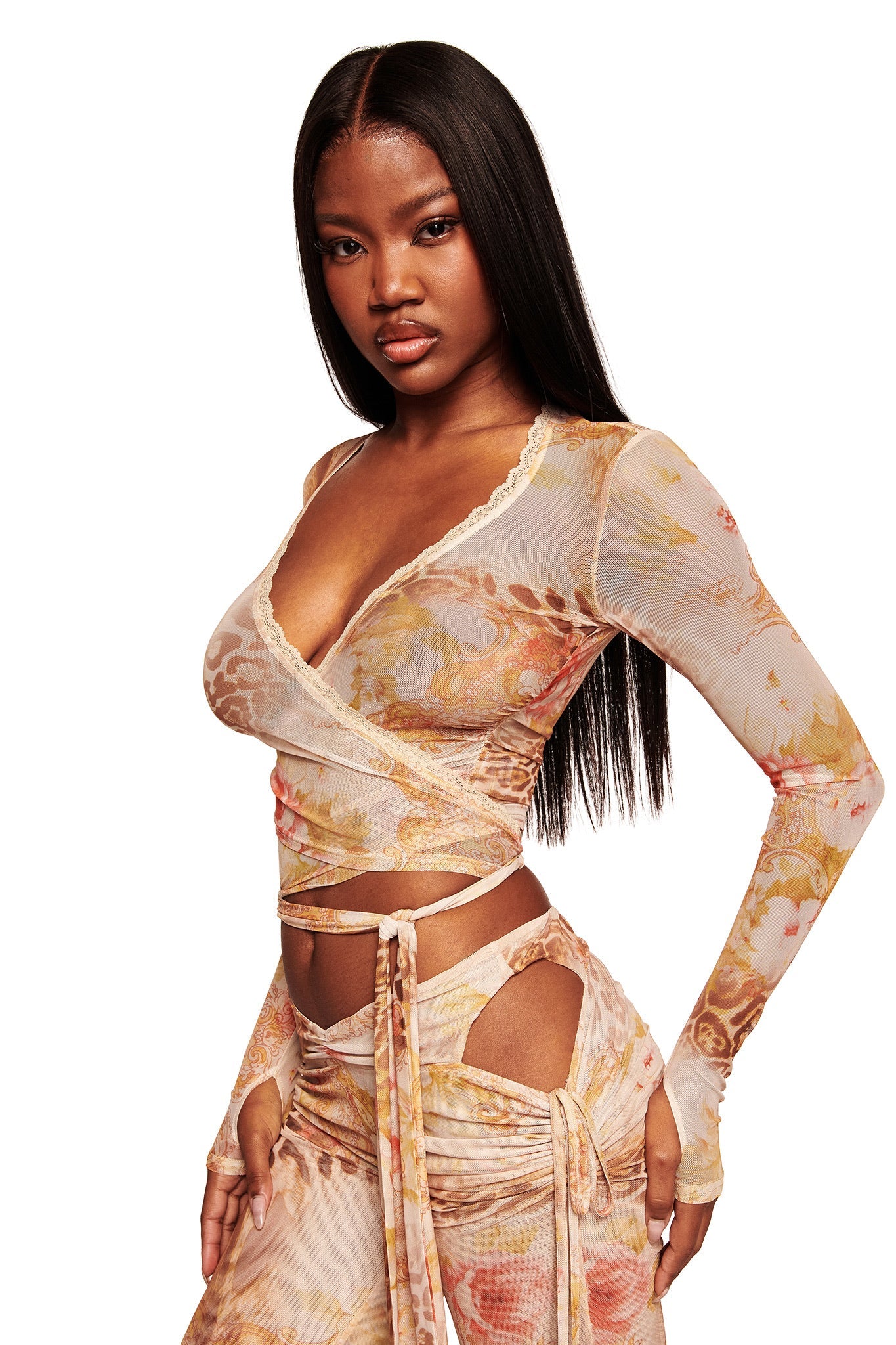 A woman with long straight hair models the I.AM.GIA ERISKA PANT in Desert Rose—mid rise mesh pants with side cutouts and a soft multicolor pattern—paired with a matching sheer wrap top, posing against a white background.