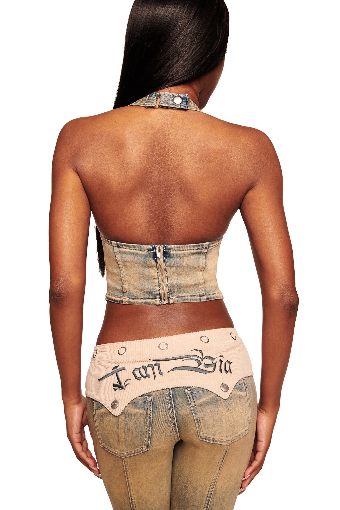 A woman with long dark hair is seen from behind wearing the I.AM.GIA TOMMIE CORSET in antique denim with matching jeans and a light belt embroidered with "I am Bia" in black script.