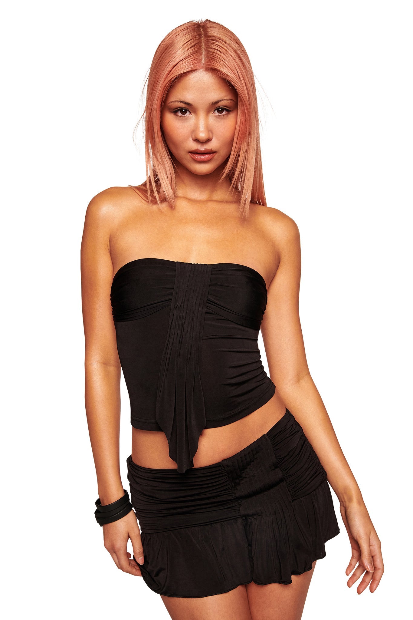 A woman with straight, pinkish-blonde hair wears the I.AM.GIA ELYSIA BOOB TUBE in black and a matching short skirt. She stands against a plain white background with a neutral expression.