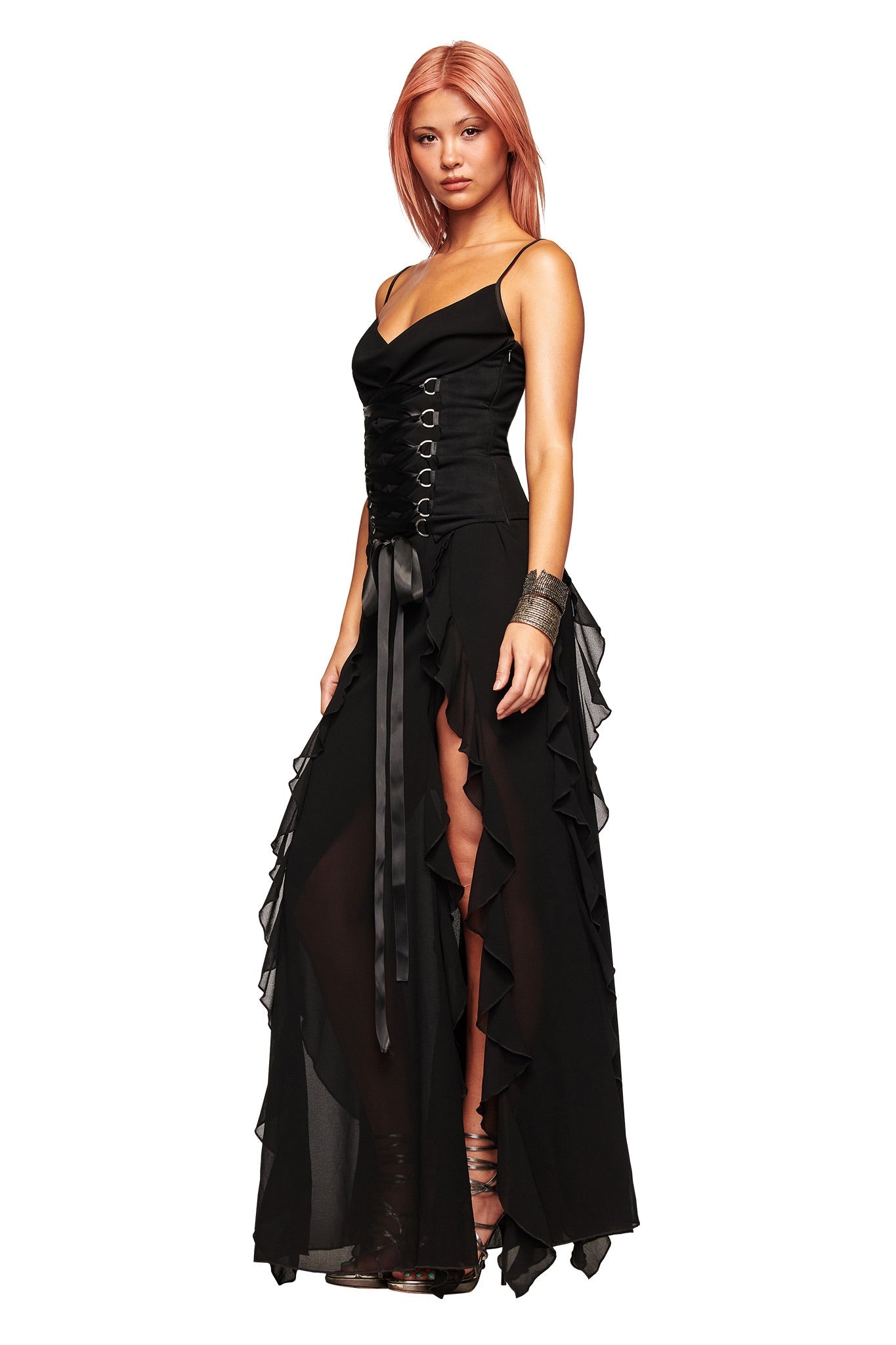 A woman with pink hair wears the I.AM.GIA MARLETTE 2.0 MAXI DRESS in black, styled with corset lacing, sheer ruffles, and black sandals, standing against a white background.