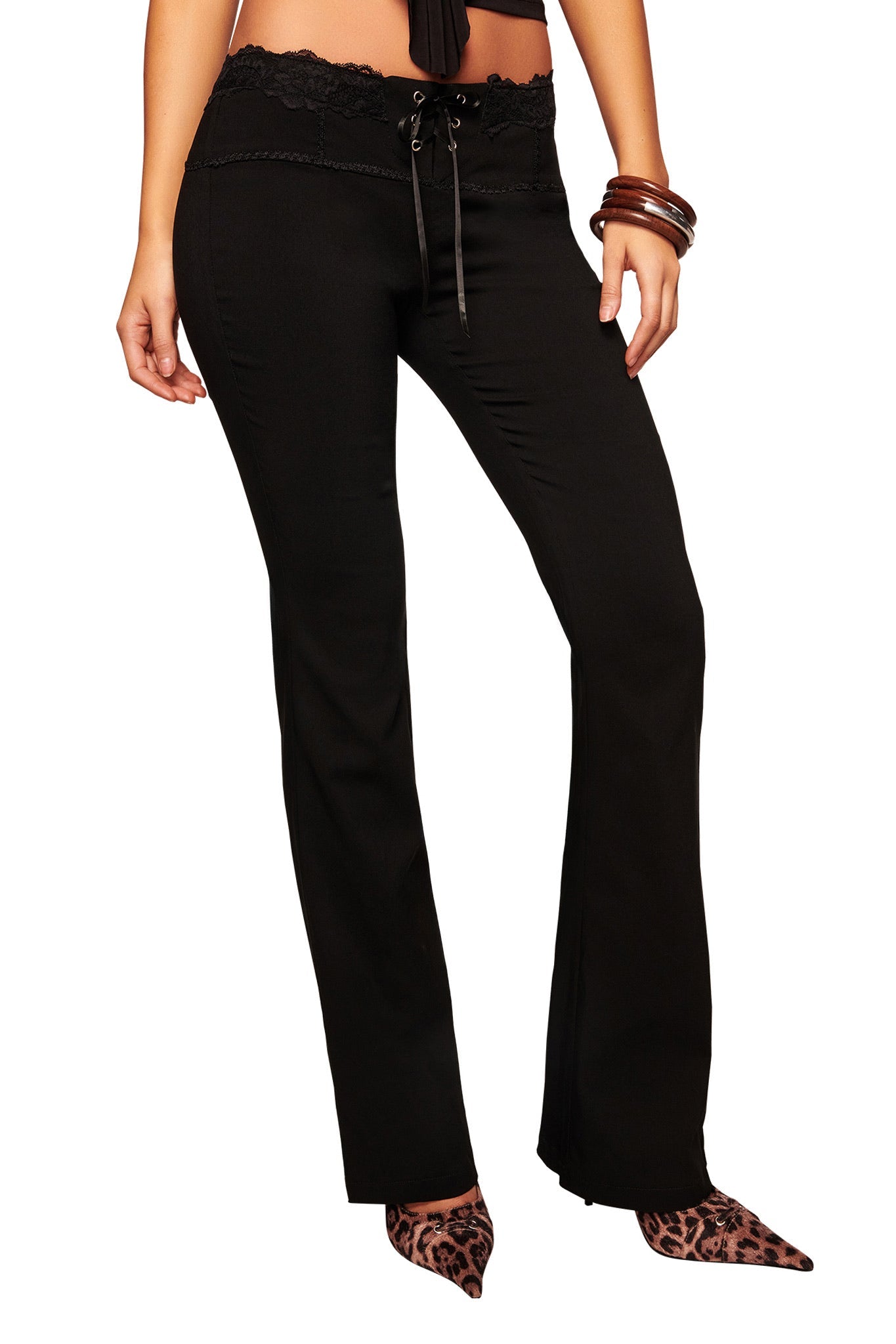 A woman wears the I.AM.GIA SERAYA PANT - BLACK, featuring a stretch woven fabric, lace waistband, and mid rise waist, styled with a black crop top, brown bangles, and leopard print pointed-toe shoes against a white background.