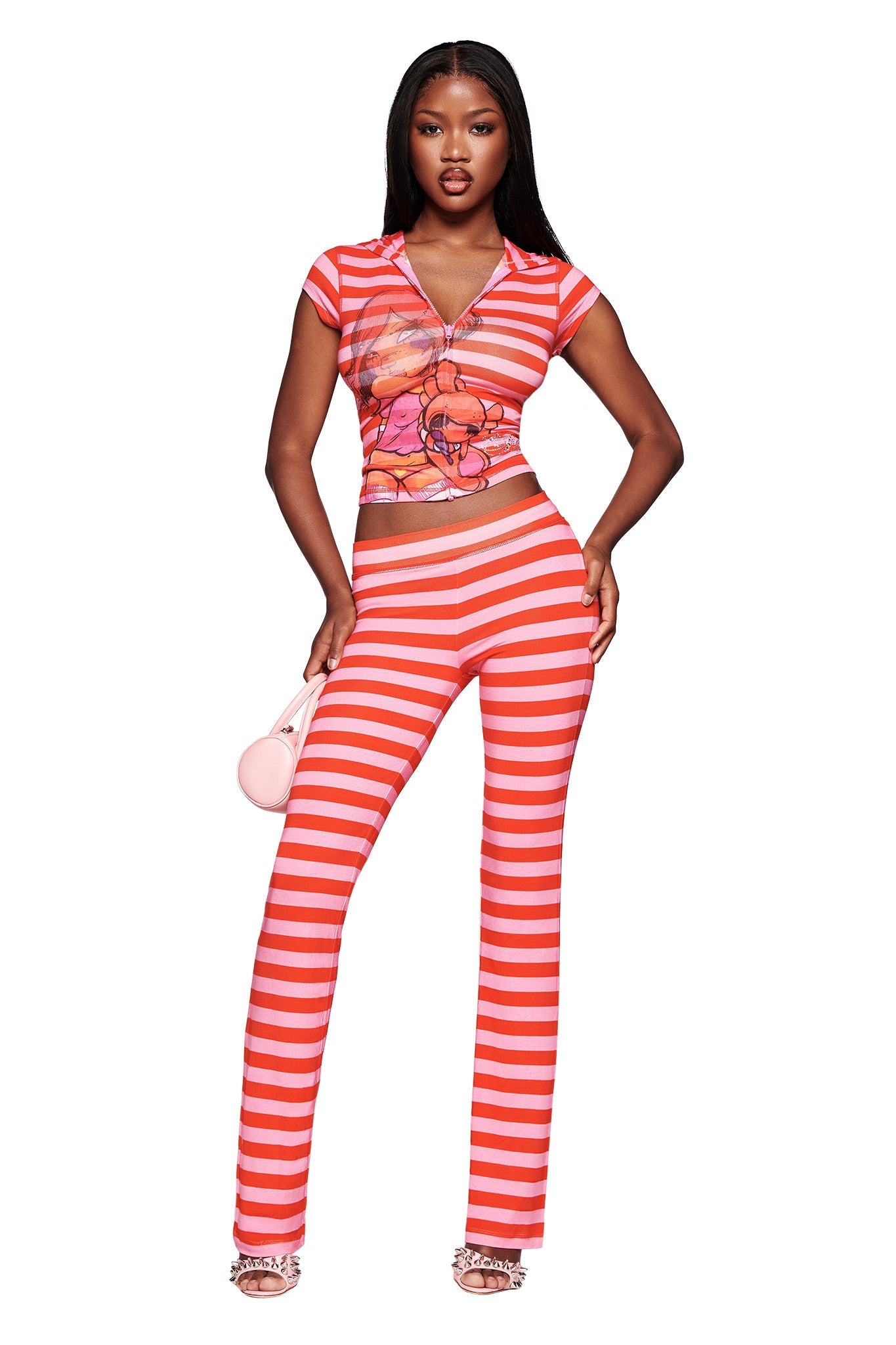 A woman poses confidently in the I.AM.GIA FAFI BLARE CAP SLEEVE HOODIE in pink stripe, paired with matching pants. She holds a small pink purse, wears open-toe heels, and stands against a white background.