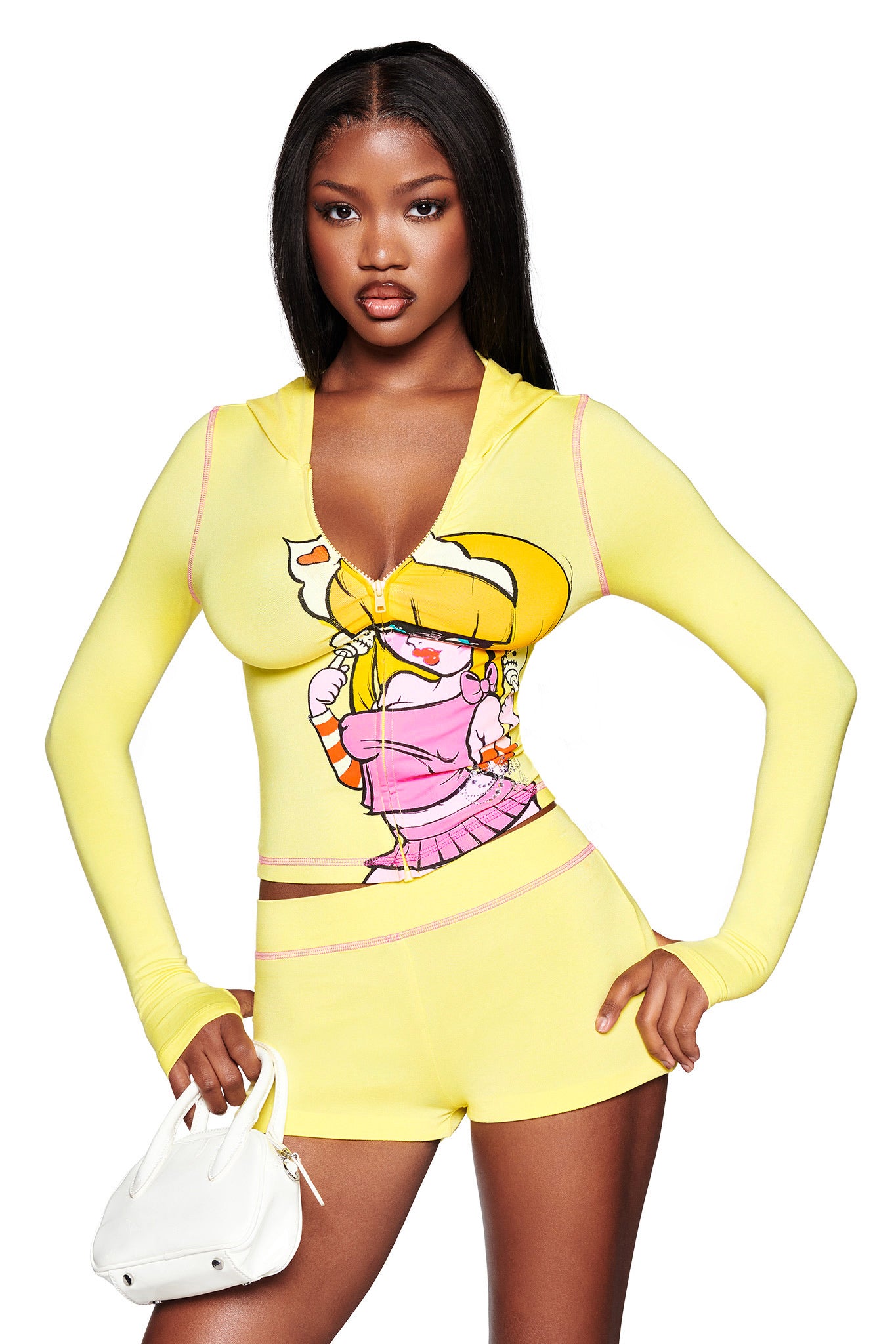 A woman wears the I.AM.GIA FAFI BLARE HOODIE - YELLOW with matching shorts, featuring a cartoon graphic, and poses confidently holding a white handbag against a white background.