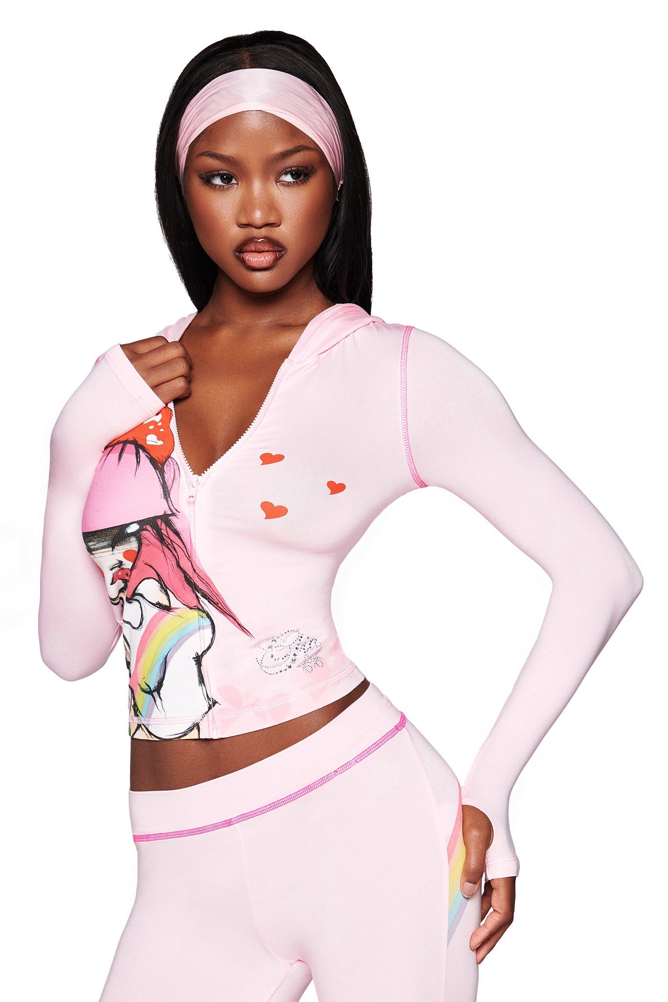 A woman poses against a white background wearing I.AM.GIA's baby pink FAFI BLARE PANT with a matching light pink Gia x Fafi hooded zip-up top featuring red hearts, rainbow art, and a headband, hand on hip.