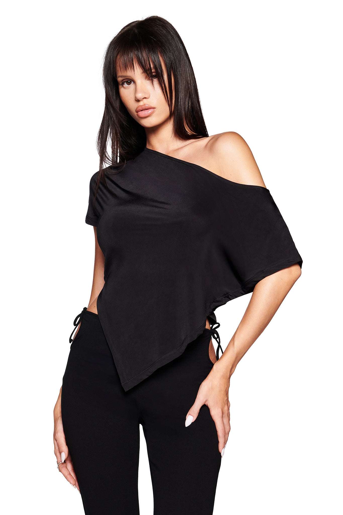 A woman wears the I.AM.GIA JOVIE TOP in black—an asymmetrical, one-shoulder top with short sleeves—paired with black bodycon pants, standing against a white background.